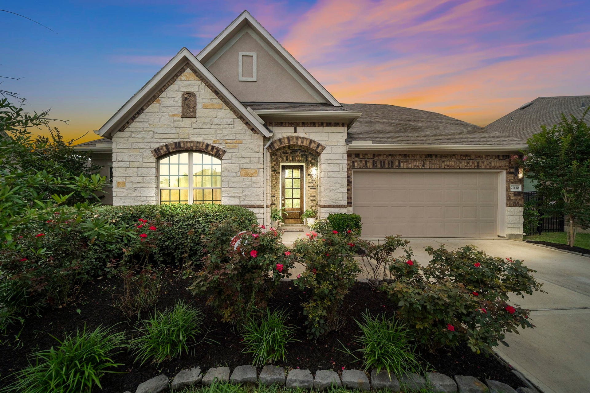 Beautiful curb appeal welcomes you home with a striking blend of stone, brick, and stucco, accented by mature landscaping and colorful blooms. The covered entry, arched architectural details, and two-car garage create a polished first impression, while the warm exterior lighting adds an inviting finishing touch.
