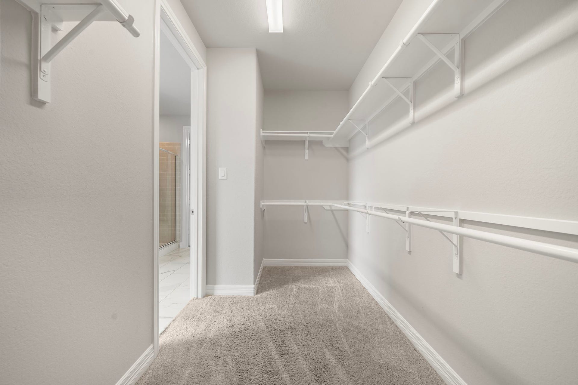 The oversized walk-in closet provides abundant hanging and shelf space along both walls, making organization easy. Soft carpeting and bright overhead lighting enhance the clean, comfortable feel, while multiple storage levels accommodate clothing, shoes, accessories, and seasonal items in a practical, well-planned layout.