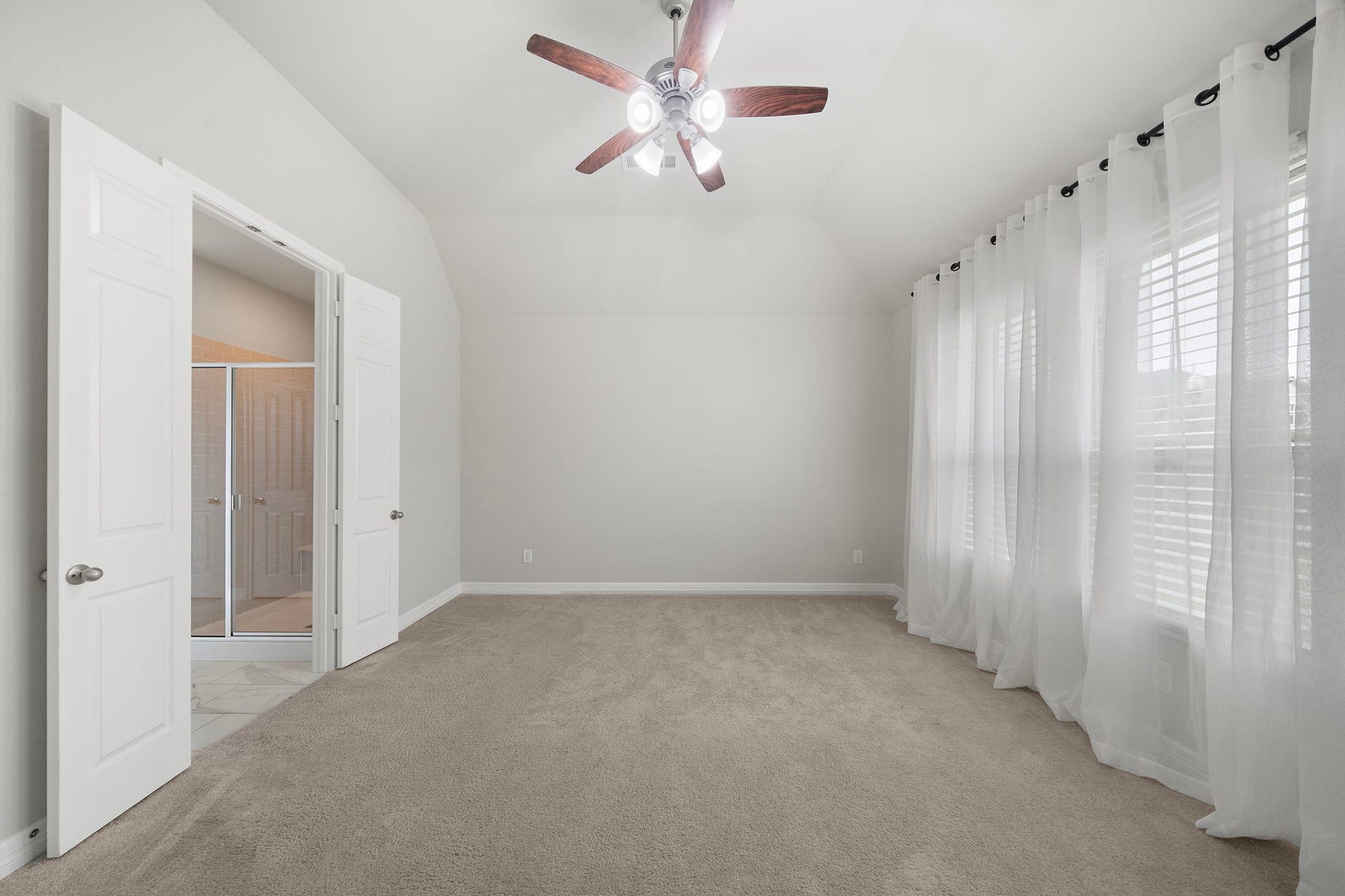 The spacious primary bedroom offers a peaceful retreat with soft carpeting, a high ceiling, ceiling fan, and a wall of windows dressed in sheer curtains. Neutral tones enhance the light and airy feel, while direct access to the adjoining bath adds comfort and convenience to this generously sized private space.