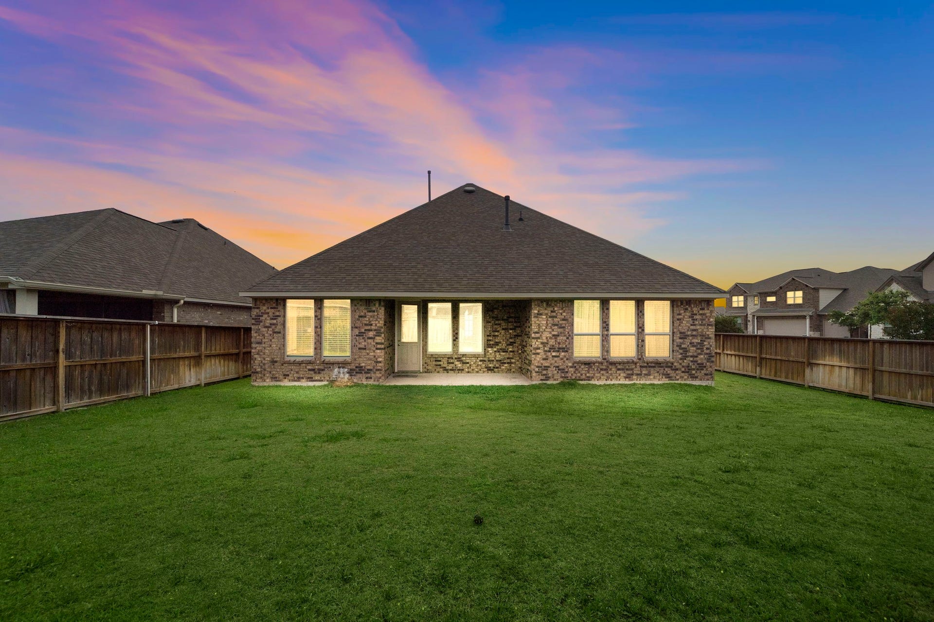 The expansive fenced backyard offers plenty of open lawn for outdoor living, recreation, gardening, or future enhancements. The brick rear exterior, covered patio area, and multiple windows create a strong connection between indoor and outdoor spaces, while the dramatic evening sky provides a beautiful backdrop for relaxing at home.