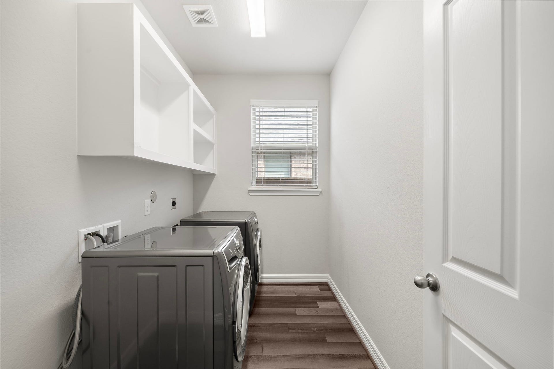 The dedicated laundry room offers a practical, well-organized space with room for a full-size washer and dryer, built-in upper shelving, bright overhead lighting, and a window that brings in natural light. Durable wood-look flooring continues the homes cohesive style while providing an attractive, easy-care surface for everyday household tasks.