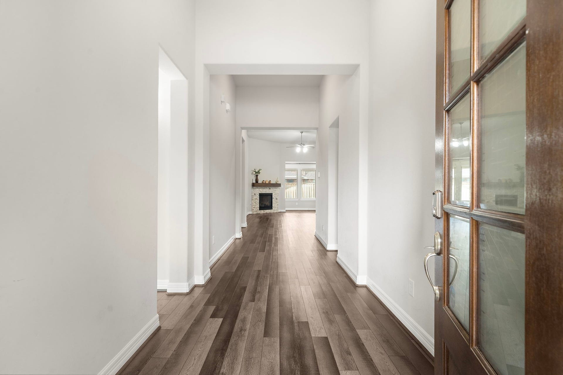 A dramatic entryway sets the tone with soaring ceilings, crisp neutral walls, and beautiful wood-look flooring extending toward the main living area. The glass-paneled front door introduces natural light, while the long, open hallway creates an impressive sense of space and an inviting transition into the heart of the home.