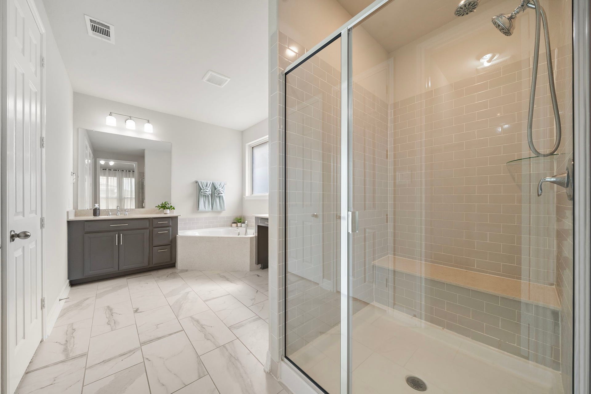The primary bath offers a spacious, spa-inspired layout with a large glass-enclosed shower, built-in bench, tiled surround, soaking tub, and extensive vanity storage. Light tile flooring and neutral finishes create a bright, relaxing environment, while the oversized mirror and multiple lighting fixtures add everyday practicality.