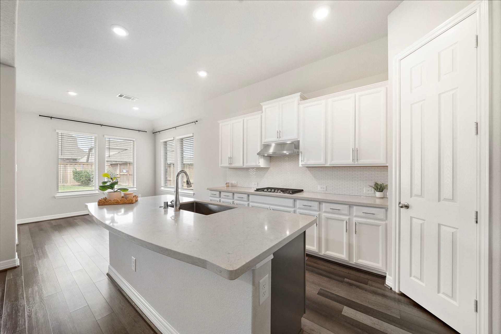 Designed with both style and function in mind, the kitchen features abundant cabinetry, a large center island with an undermount sink, sleek countertops, and a striking stainless steel vent hood above the gas cooktop. A classic tile backsplash, recessed lighting, and natural light from surrounding windows complete this bright, polished space.