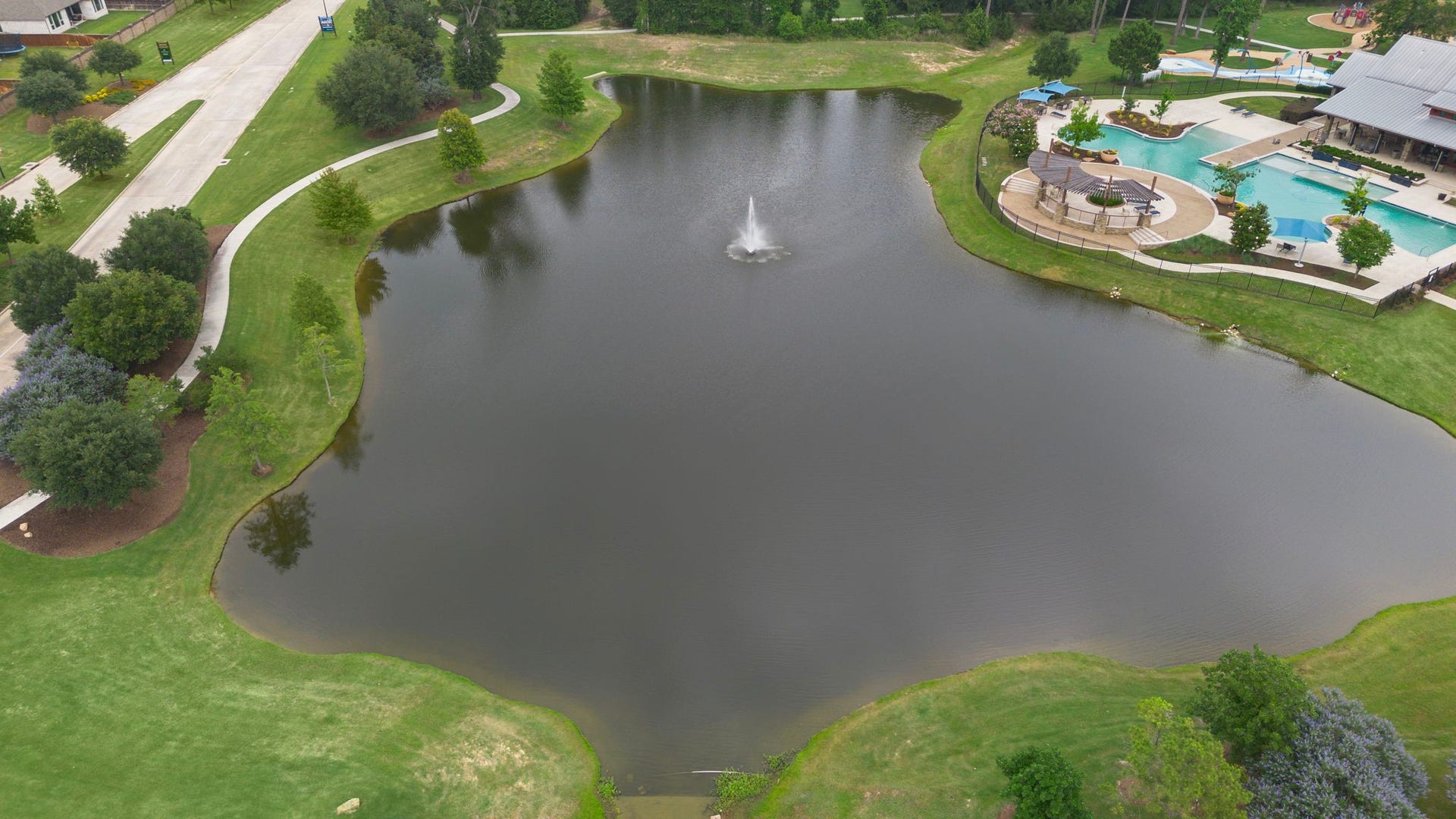 A scenic community pond creates a peaceful natural centerpiece surrounded by manicured lawns, mature landscaping, and winding walking paths. A central fountain adds visual interest, while the nearby resort-style pool and recreational amenities complement the setting, offering residents a beautiful environment for walking, relaxing, and enjoying the outdoors.