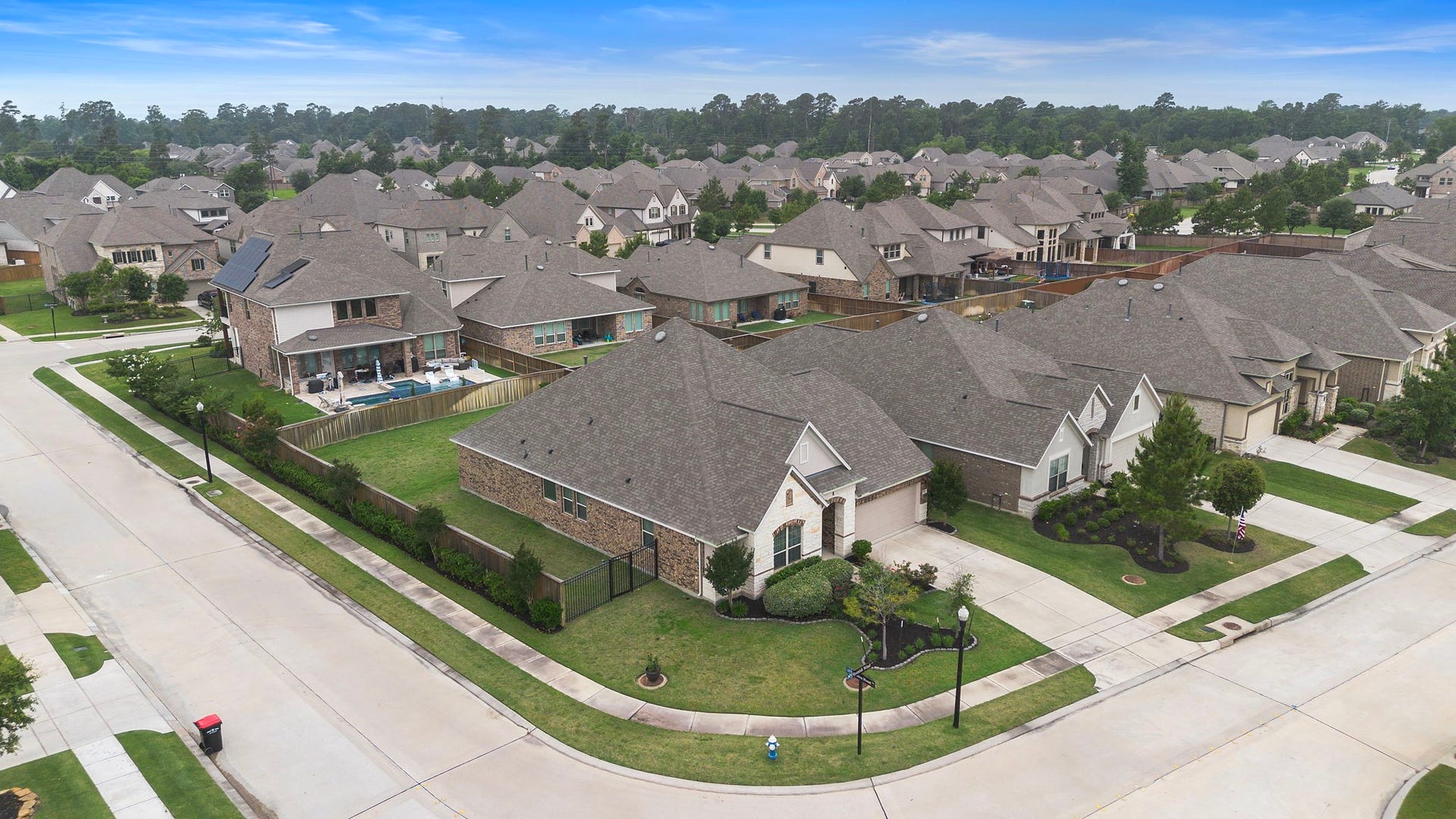 This aerial perspective provides a broader look at the property and surrounding neighborhood, showcasing the homes substantial footprint, fenced backyard, landscaped front exterior, and nearby residences. Curving streets, sidewalks, mature trees, and thoughtfully maintained homes contribute to the inviting character of the area.