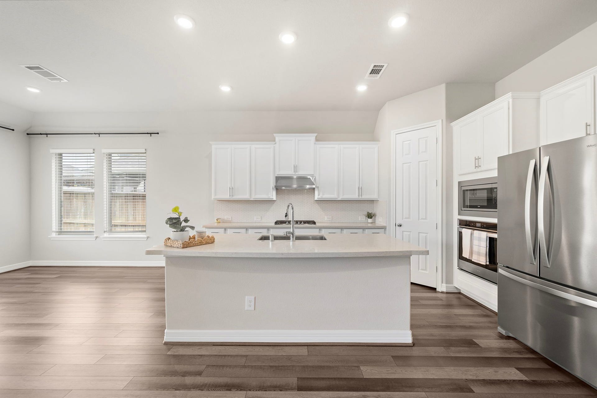 Clean lines and thoughtful design define this spacious kitchen, where a generous center island provides excellent prep space and casual seating. White cabinetry, stainless steel appliances, recessed lighting, and neutral countertops create a timeless look, while multiple windows bring in natural light and enhance the open, airy atmosphere.