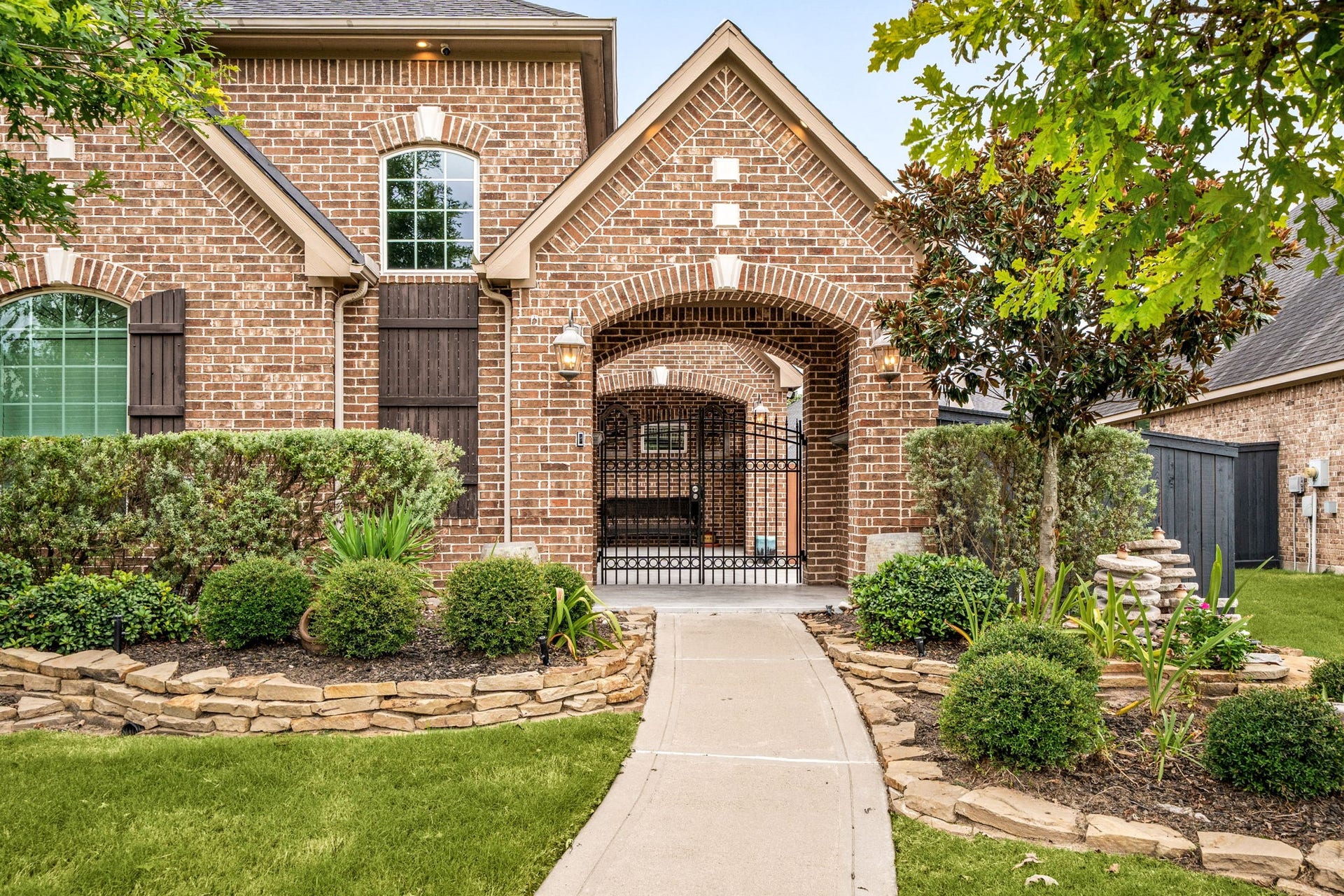 A wrought-iron gated courtyard welcomes you home, framed by an arched brick entry, mature landscaping, and a stone-lined walkway  a custom feature that adds privacy and curb appeal.