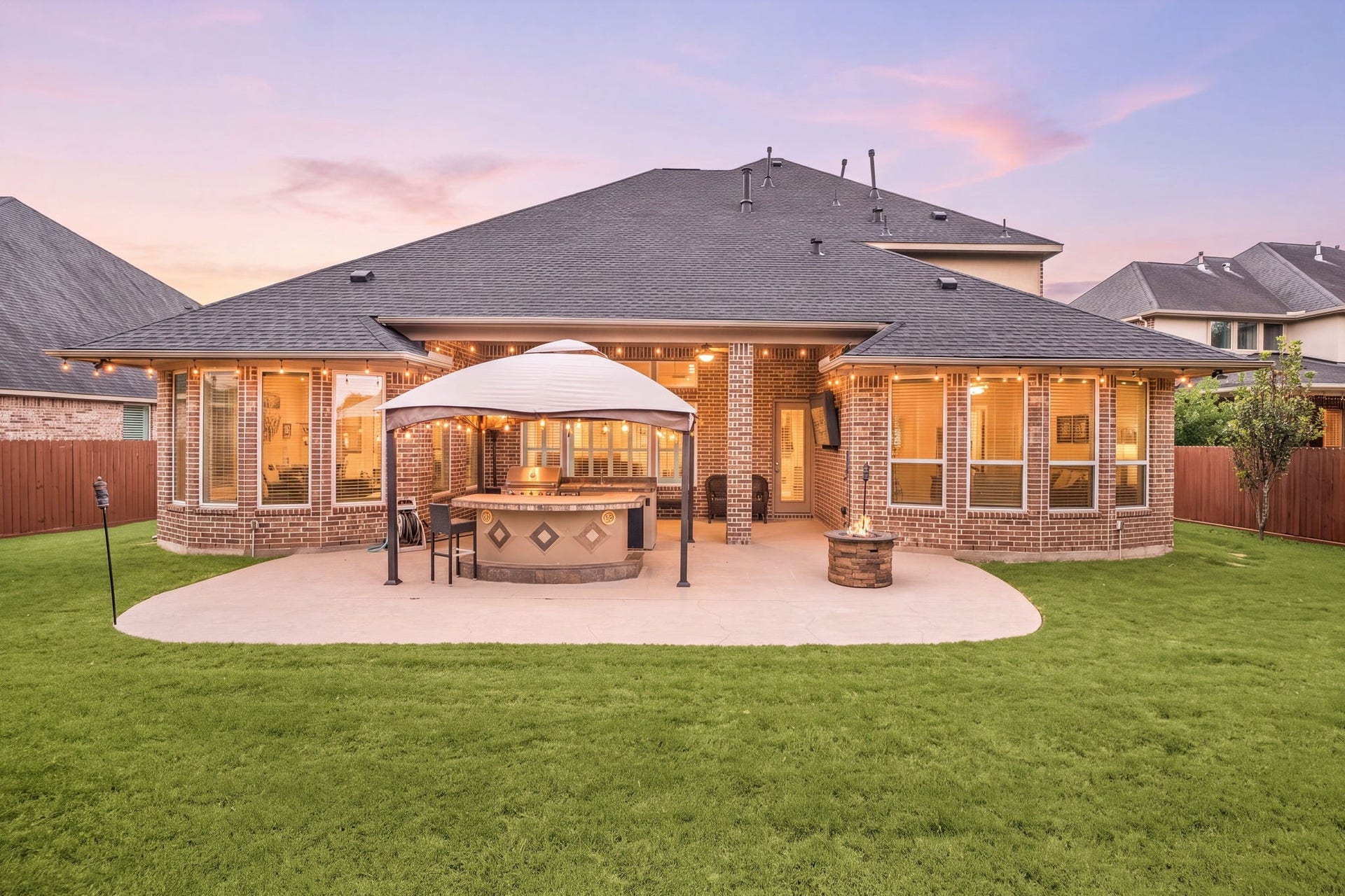 An entertainer's dream at twilight, the custom outdoor kitchen and covered patio glow under string lighting, framed by a spacious, fully fenced backyard.