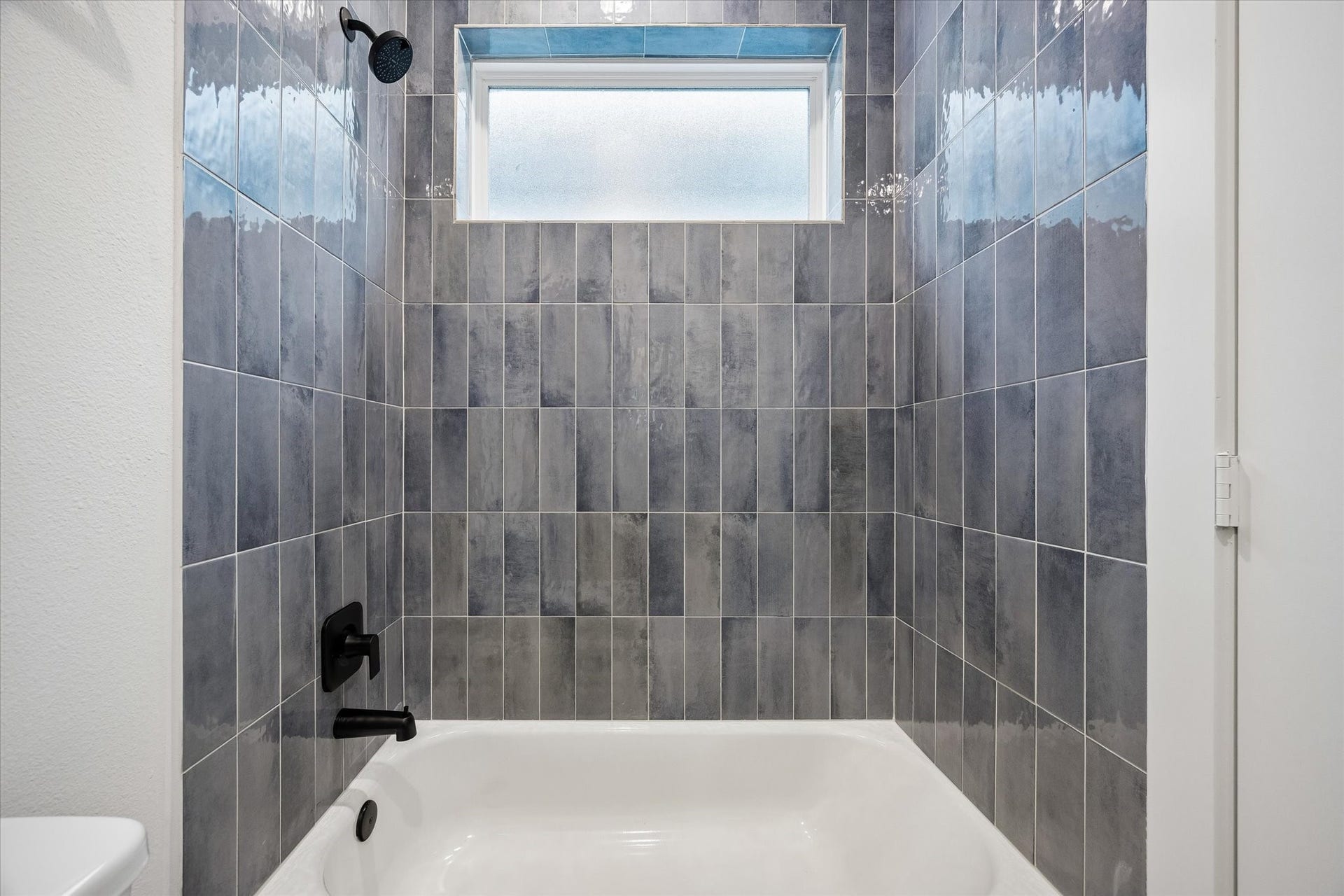 Modern bathroom with sleek gray tile walls, featuring a pristine bathtub and stylish black fixtures, illuminated by a frosted window for natural light.