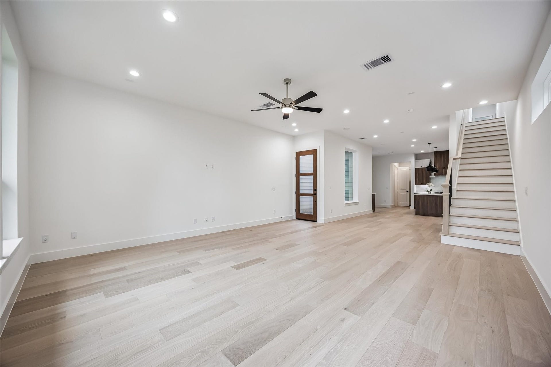 Spacious and bright living area featuring elegant wood flooring, recessed lighting, and a modern ceiling fan, seamlessly leading to a contemporary kitchen and staircase.