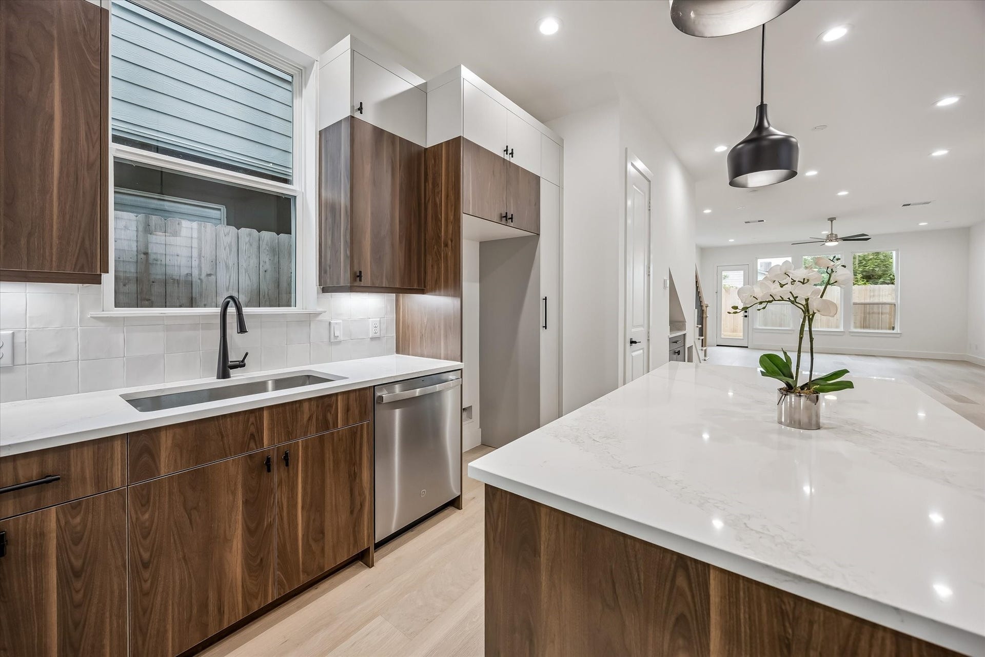 Sleek modern kitchen with rich wood cabinetry, a spacious island with elegant countertops, and stainless steel appliances, opening to a bright living area with large windows.
