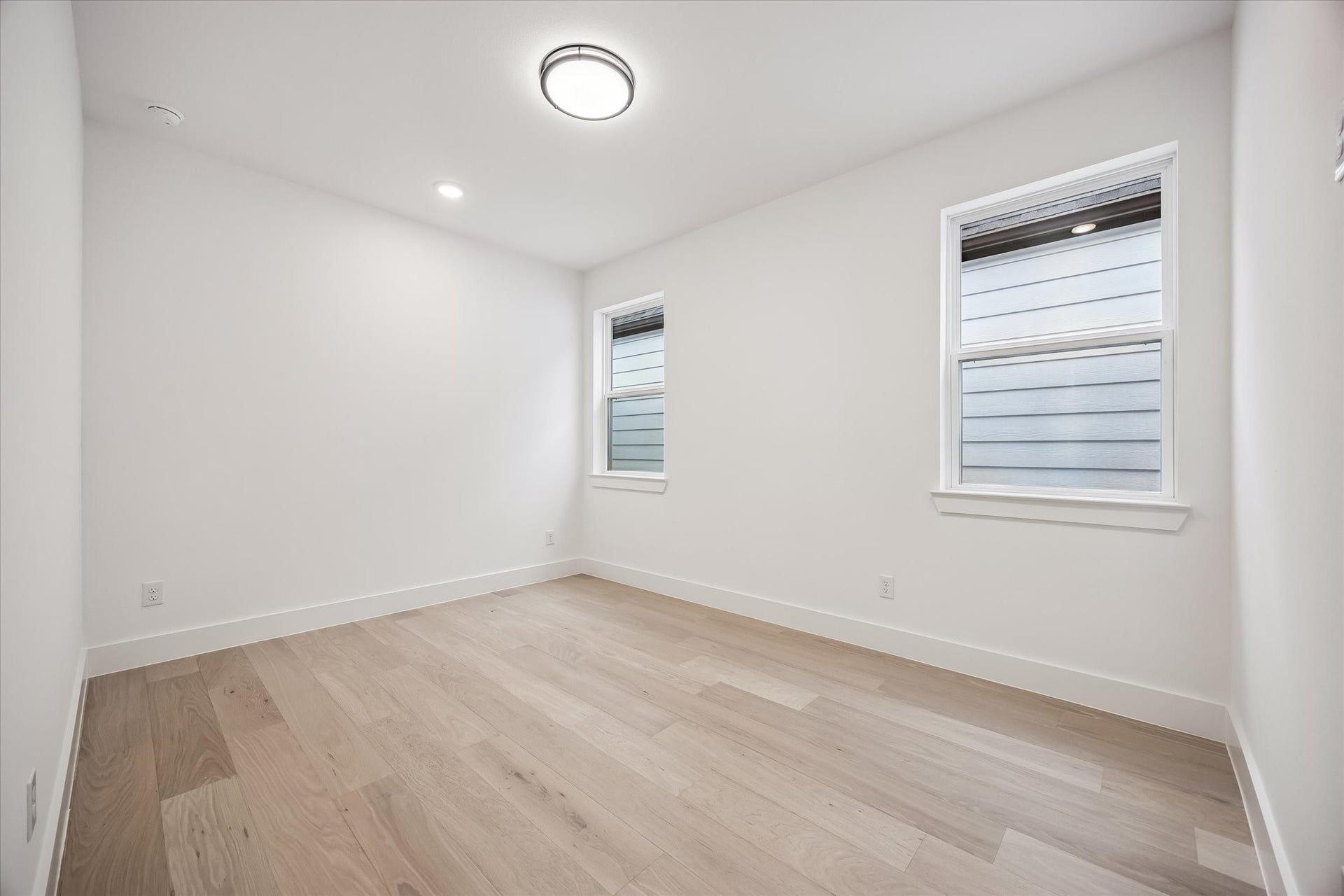 Bright and airy room featuring fresh white walls, light hardwood flooring, and ample natural light through dual windows, perfect for a cozy bedroom or home office.