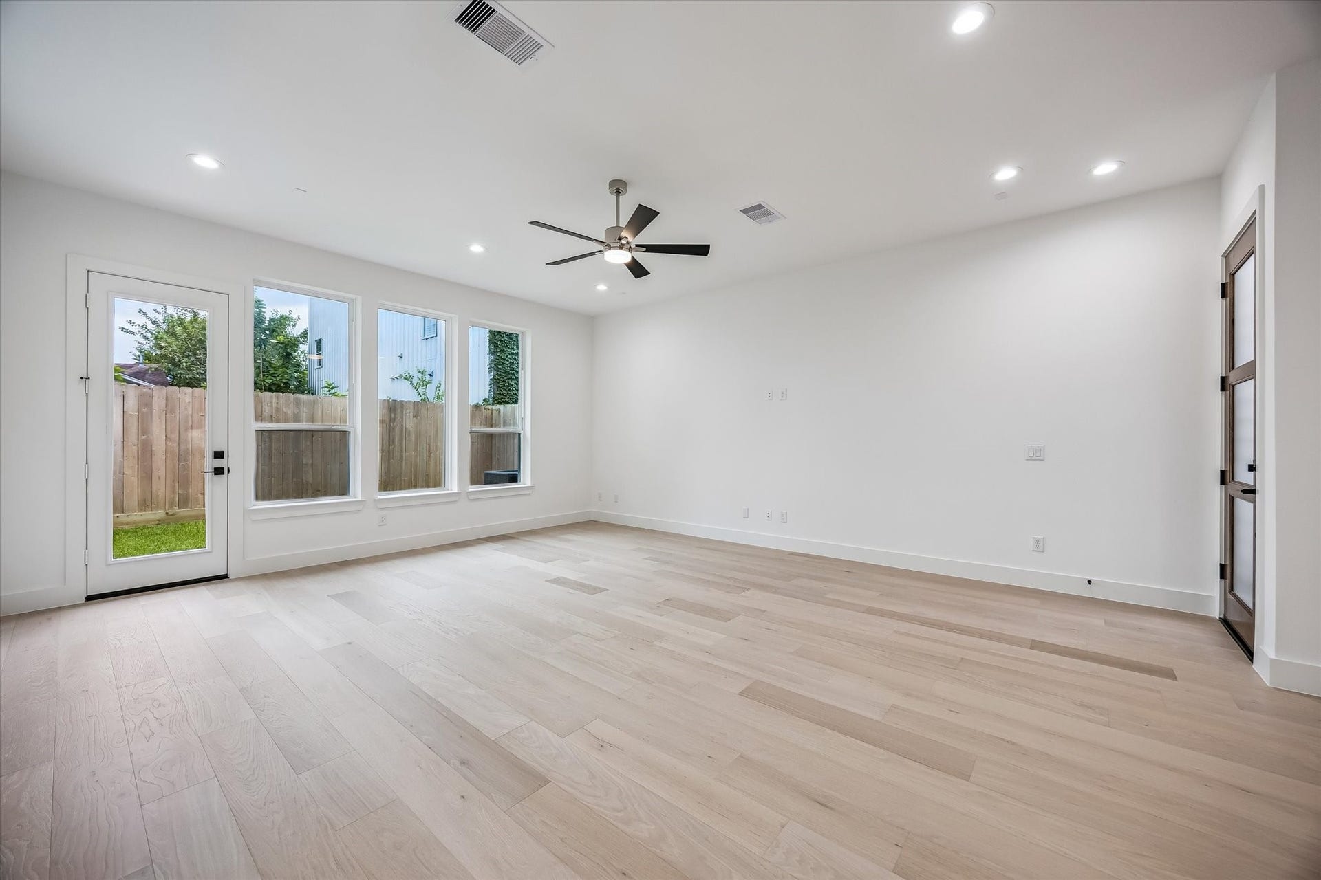 Spacious living area with light wood flooring, abundant natural light from large windows, and a modern ceiling fan, creating an inviting atmosphere.