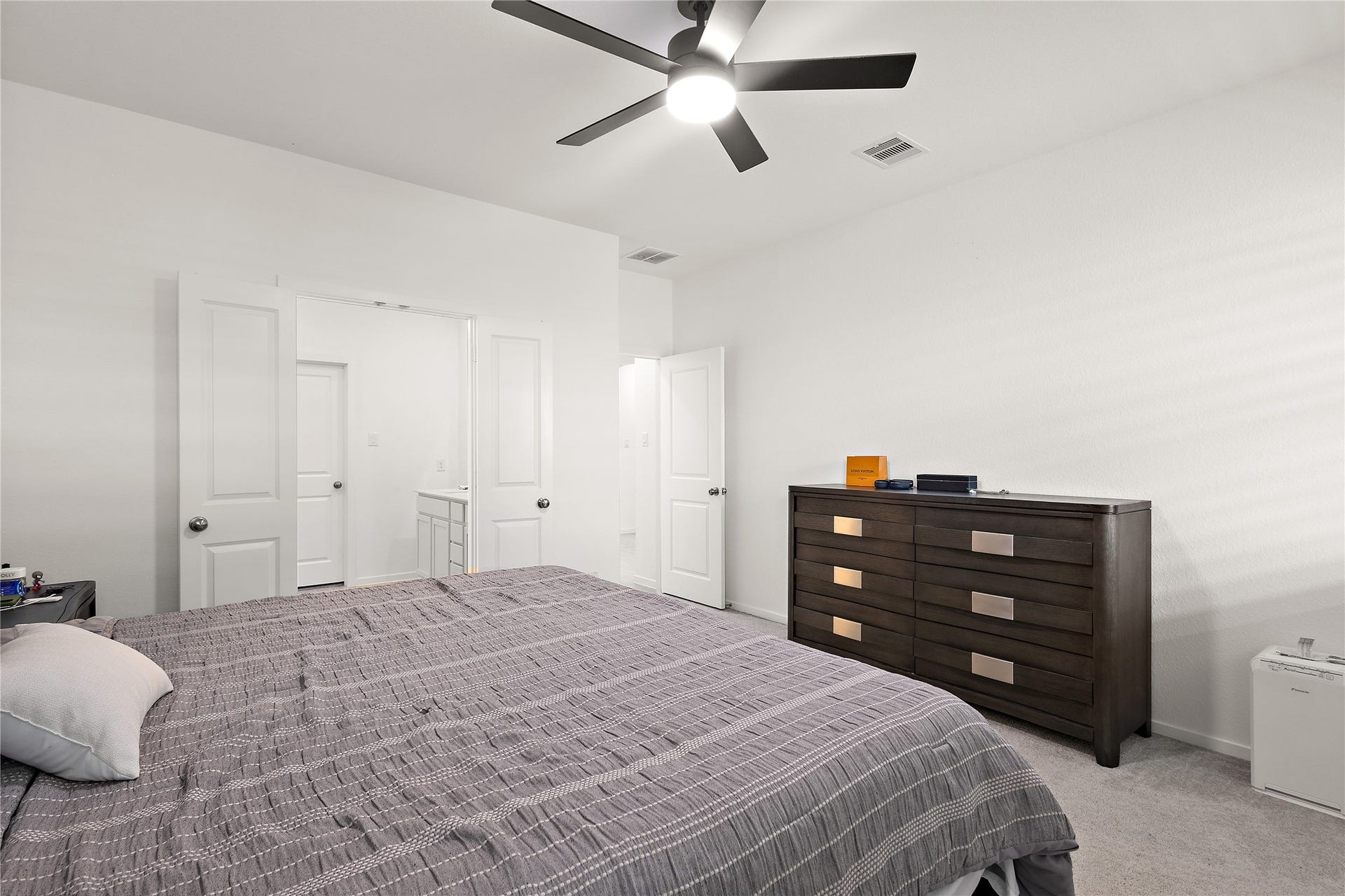 3414 Eden Valley Lane, Rosenberg, TX 77471 This bedroom features a modern ceiling fan, neutral walls, and a spacious layout. It includes a large bed with a dark wood dresser, and an ensuite bathroom is accessible through a door on the left.