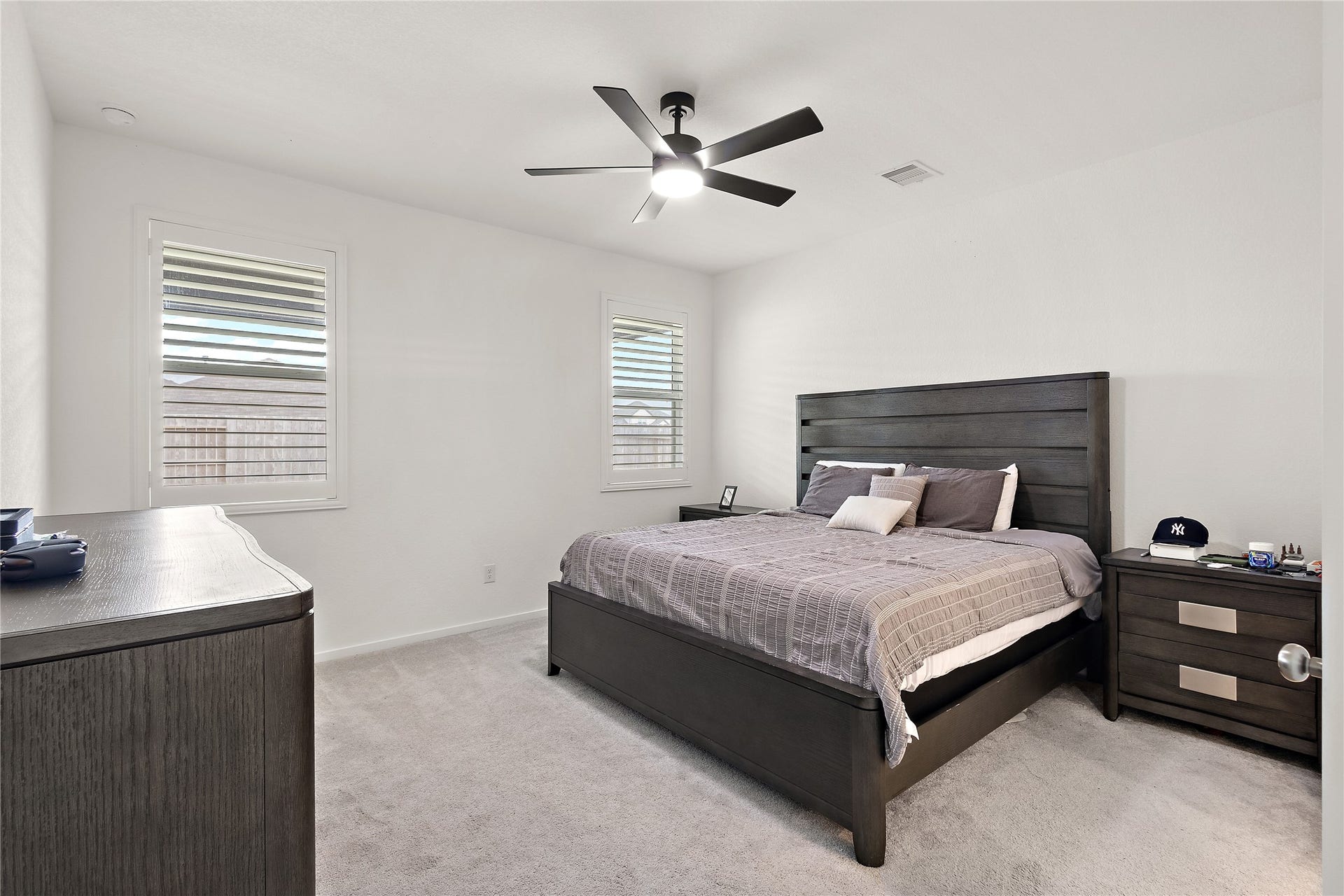 3414 Eden Valley Lane, Rosenberg, TX 77471 This bedroom features a modern, minimalist design with a dark wood bed frame and matching nightstands. It has neutral-colored walls, ample natural light from two windows, and a ceiling fan for comfort.