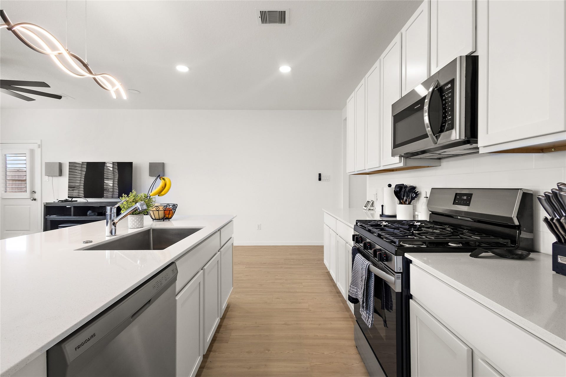 3414 Eden Valley Lane, Rosenberg, TX 77471 Modern kitchen with sleek white cabinetry, stainless steel appliances, and a large island with a sink. The space features recessed lighting and opens into a living area with light wood flooring, creating a bright and spacious atmosphere.