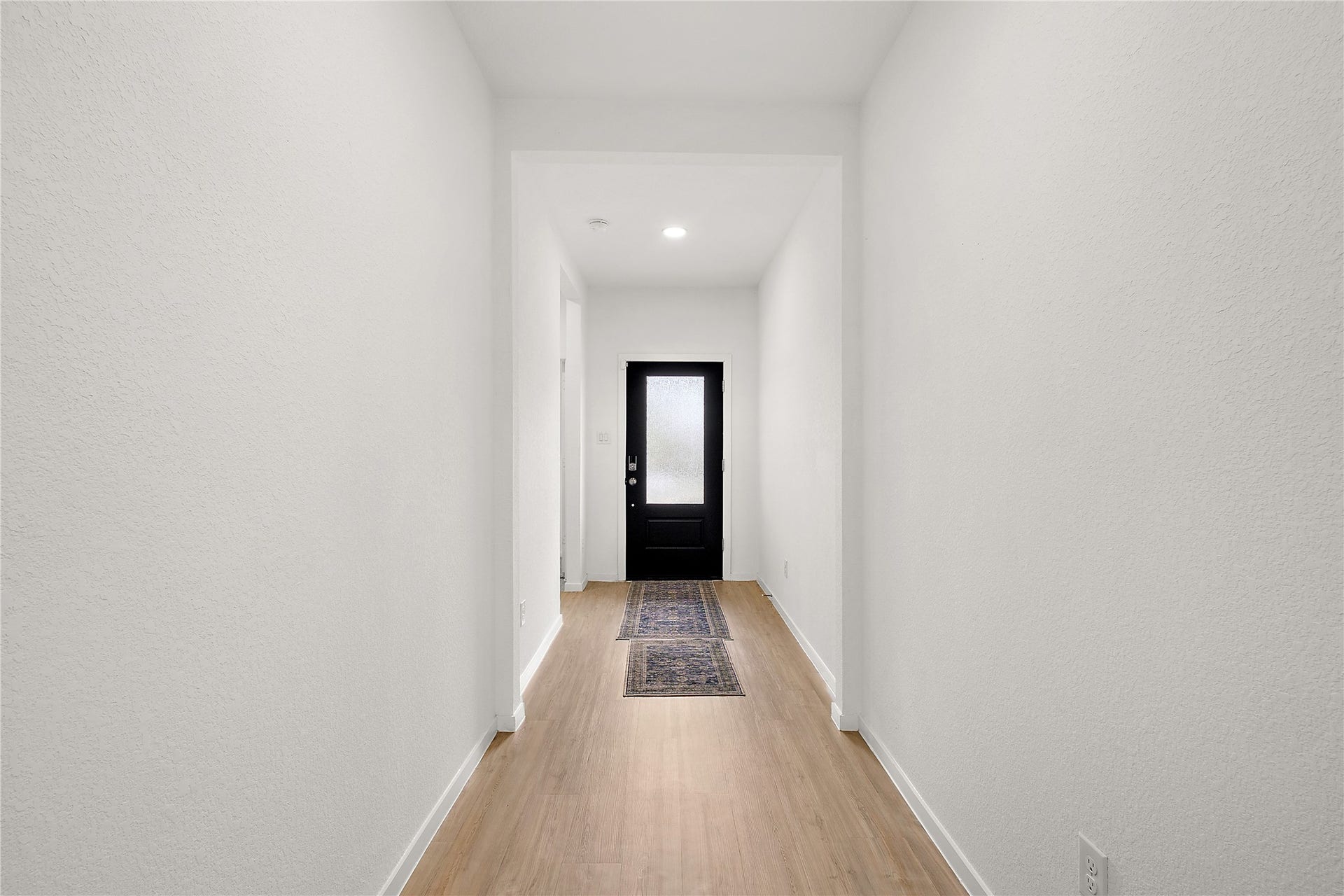 3414 Eden Valley Lane, Rosenberg, TX 77471 This photo shows a bright, minimalistic hallway with light wood flooring and white walls. It features a black front door with frosted glass, creating a modern and welcoming entryway. A narrow rug adds a subtle touch of warmth.