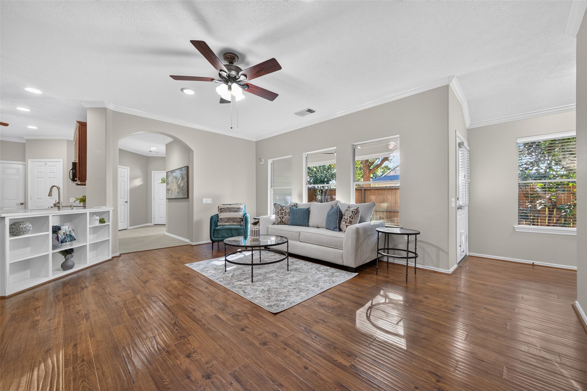 This inviting living room features a soothing ceiling fan and a wall of windows that fills the space with natural light. The open flow to the kitchen creates a bright, connected atmosphere with plenty of room to relax and unwind.