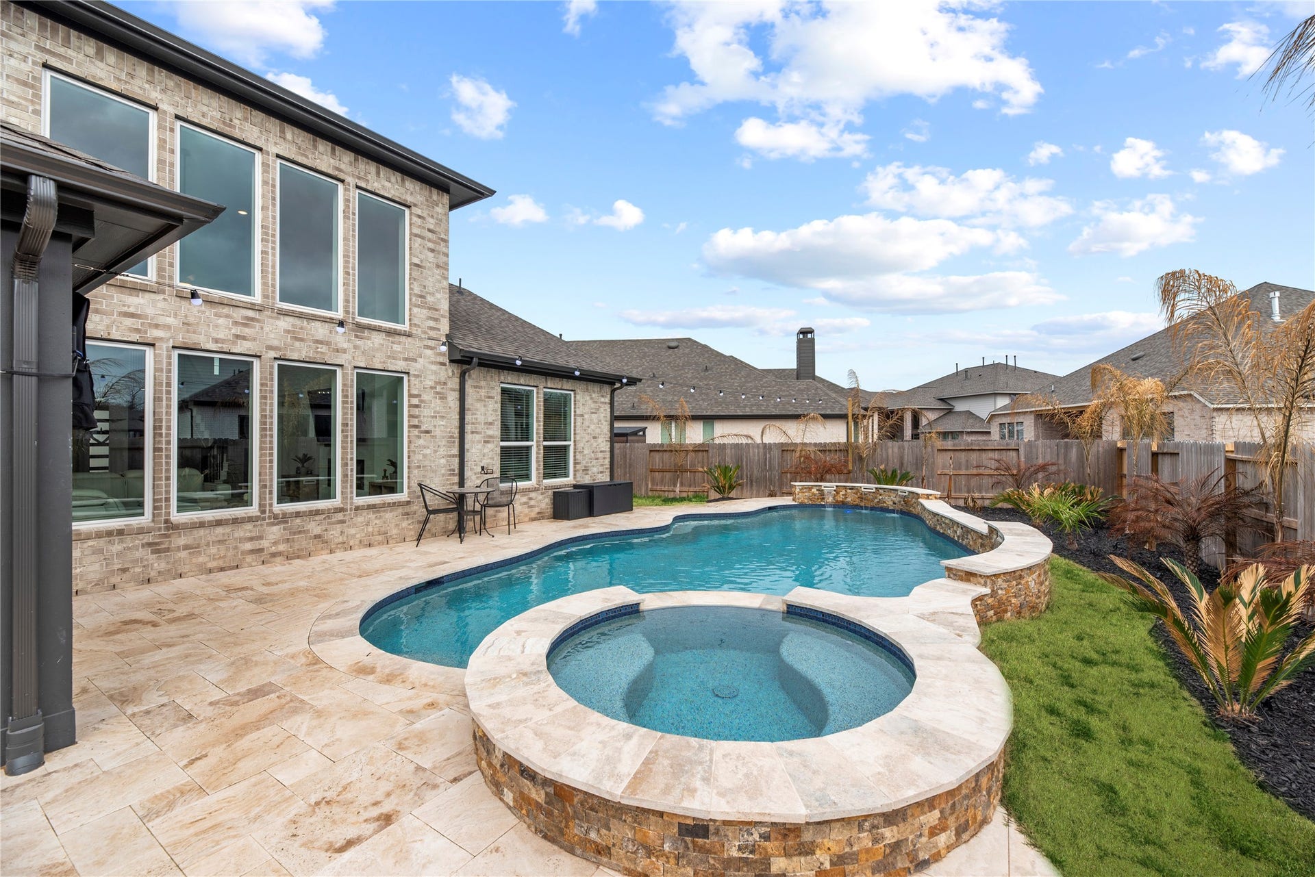 From this angle, the pool highlights the integrated raised spa, finished in stacked natural stone and complemented by travertine-style pavers that coordinate with the surrounding deck.