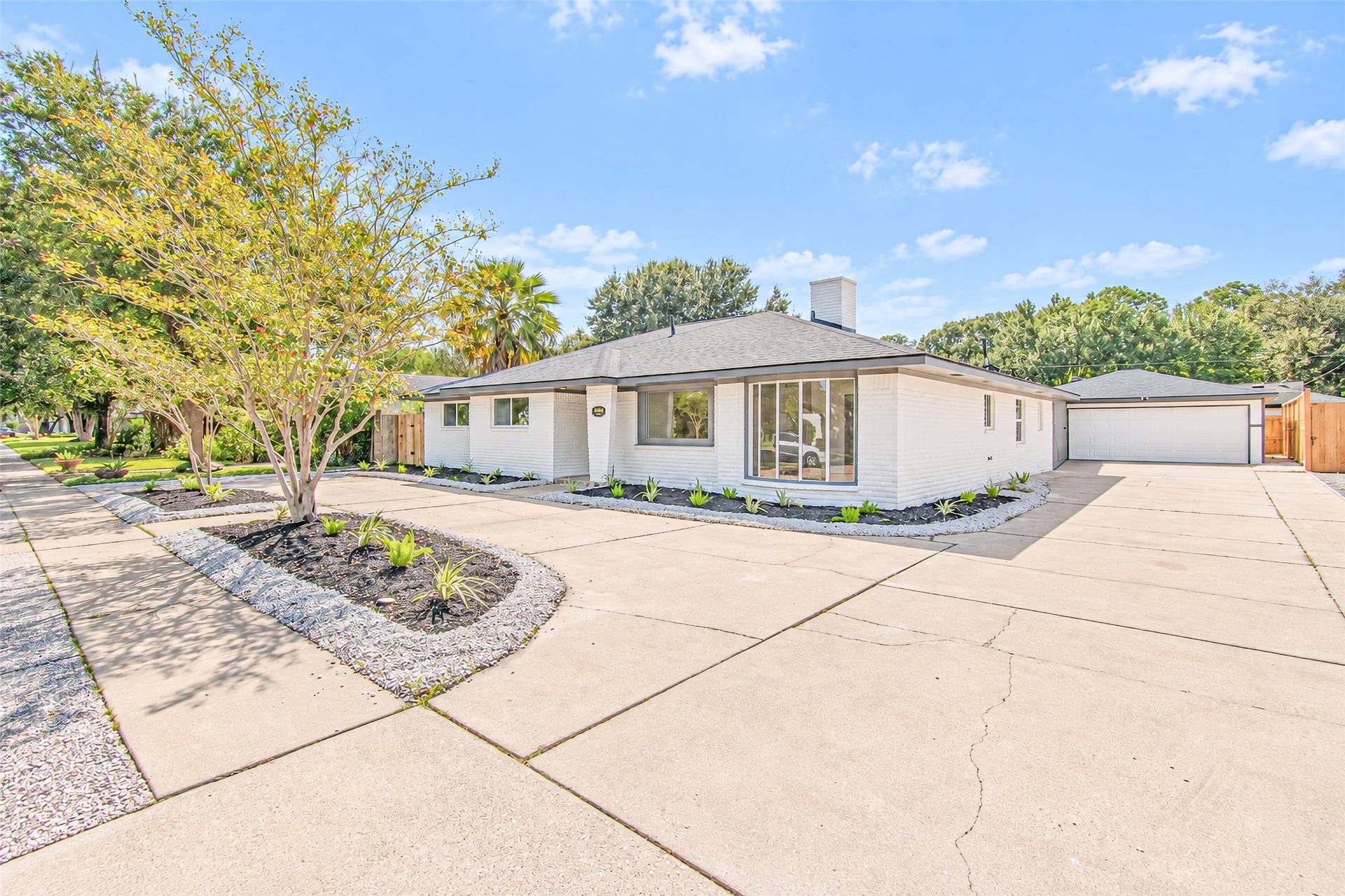 Charming single-story home with a modern white exterior, expansive driveway, and beautifully maintained landscaping, perfectly nestled in a tranquil neighborhood setting.
