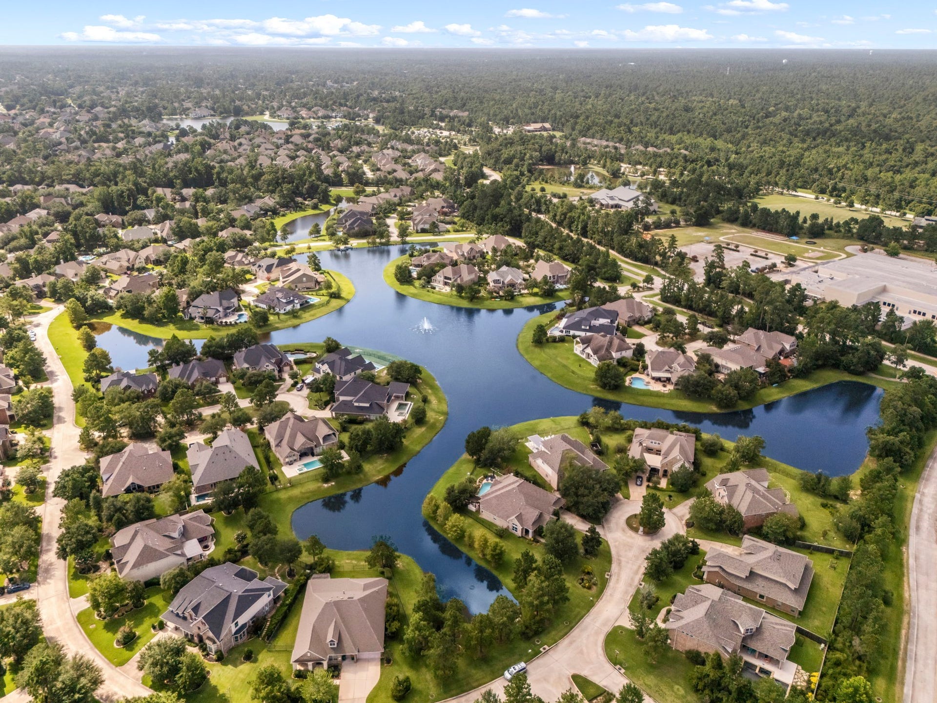 195 Oarwood Place, The Woodlands, TX 77389 This stunning high-angle aerial view showcases a breathtaking master-planned neighborhood wrapped completely around a sprawling, pristine community lake with active water fountains. Properties enjoy premium waterfront positioning, premium cul-de-sac streets, and deep setbacks flanked by manicured green lawns and mature tree lines. This peaceful enclave offers residents an unmatched resort-style daily backdrop with private loop streets and immediate proximity to walking paths and nature.