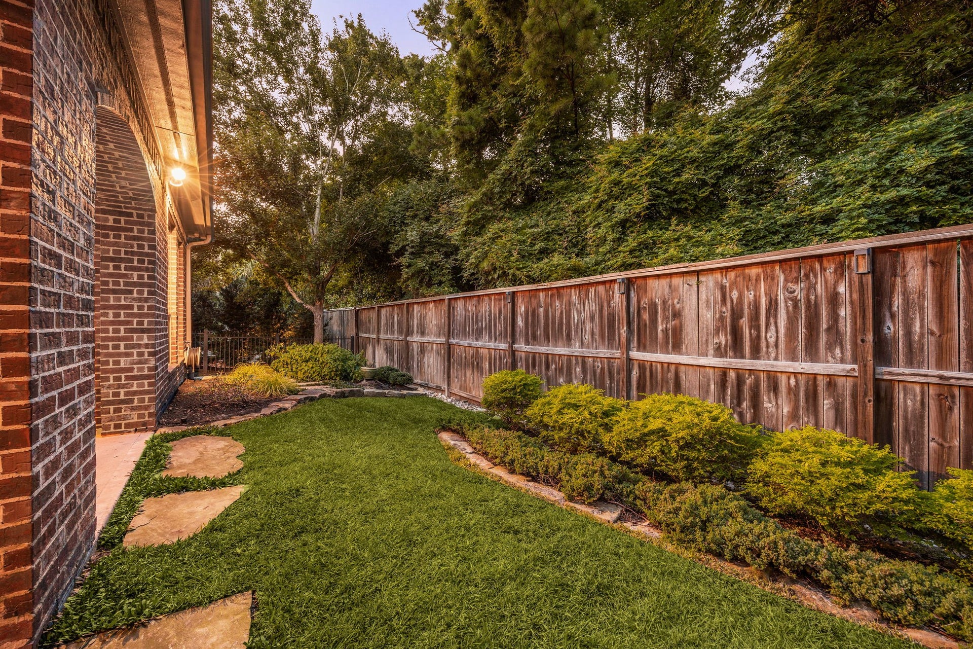 195 Oarwood Place, The Woodlands, TX 77389 This side perspective highlights the exceptional privacy and pristine landscaping of the propertys fully fenced backyard. A beautifully manicured lawn is lined with structured evergreen shrub beds and custom natural stone landscape edging, all bordered by a sturdy privacy fence and a backdrop of mature, towering trees. Integrated flagstone stepping paths trace the side of the timeless brick estate, creating a serene, highly usable outdoor escape.