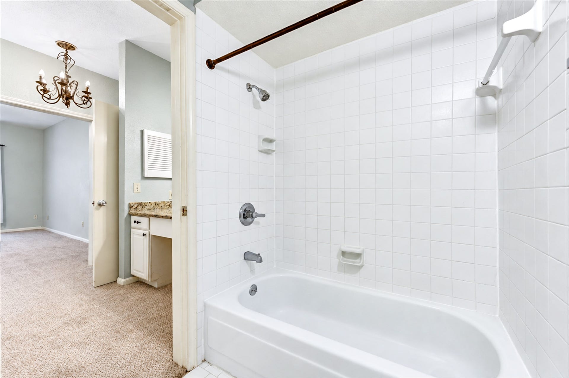 Bright bathroom features a clean white tiled shower-tub combo with convenient built-in shelving, adjacent to a cozy carpeted bedroom with elegant chandelier.