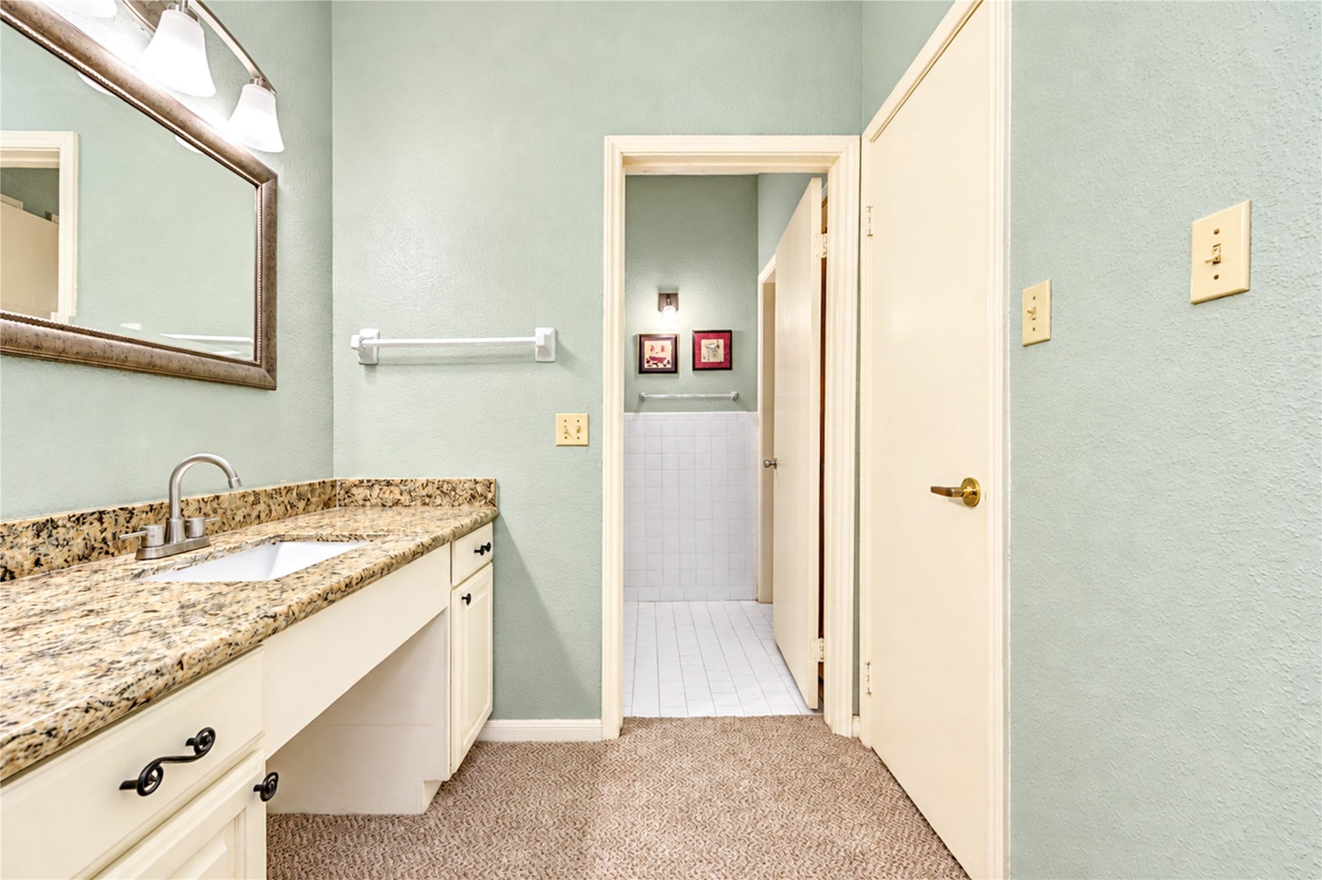 Elegant bathroom with a granite countertop and stylish lighting, leading into a cozy, carpeted bedroom with natural light and a ceiling fan.