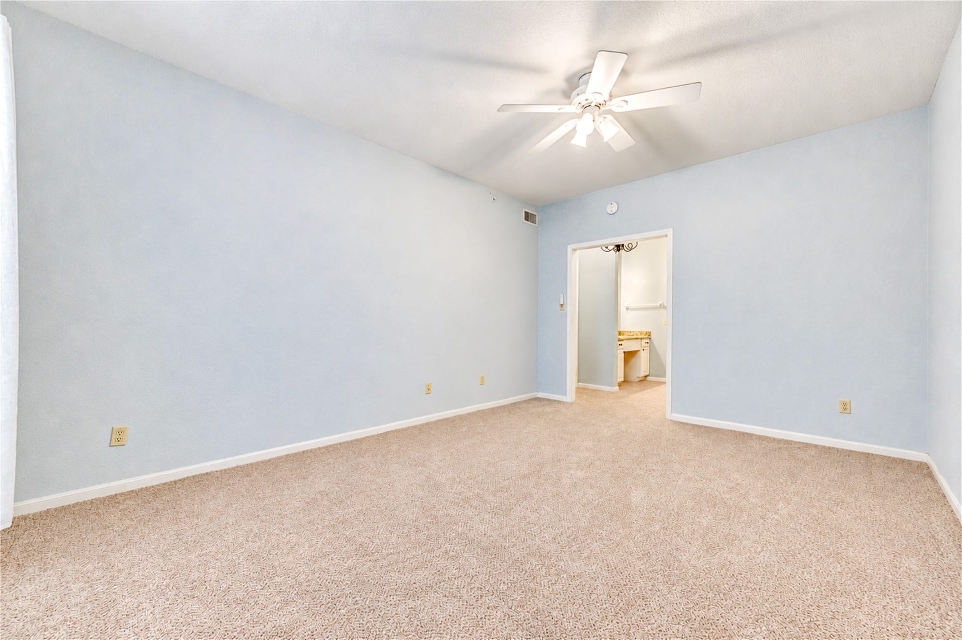 Spacious bedroom featuring soft blue walls, cozy carpeted flooring, and a ceiling fan for comfort, leading to an adjacent bathroom.