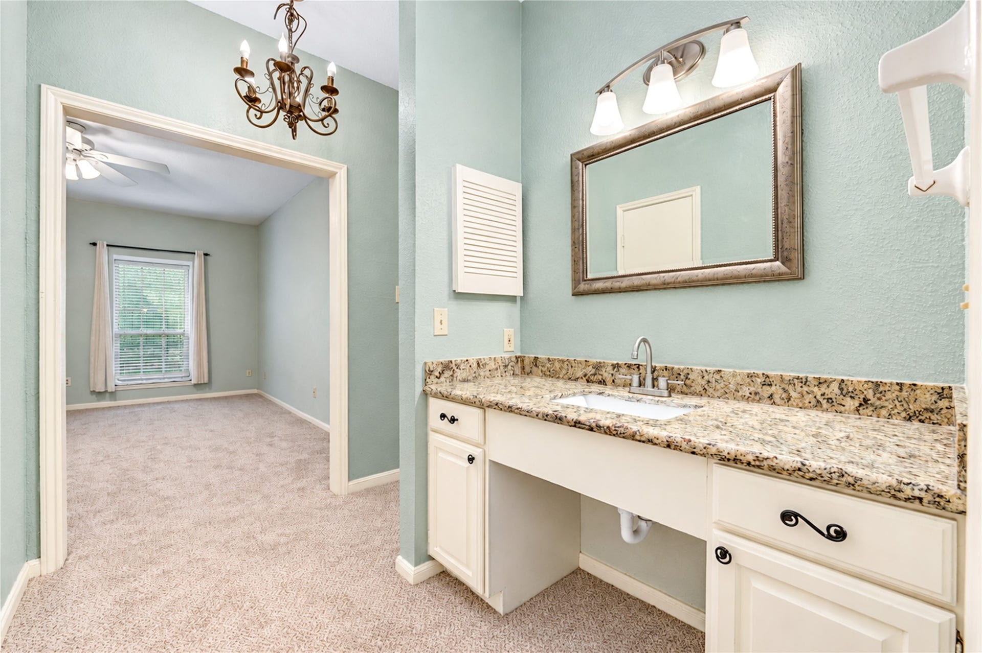 Bright bathroom features a clean white tiled shower-tub combo with convenient built-in shelving, adjacent to a cozy carpeted bedroom with elegant chandelier.