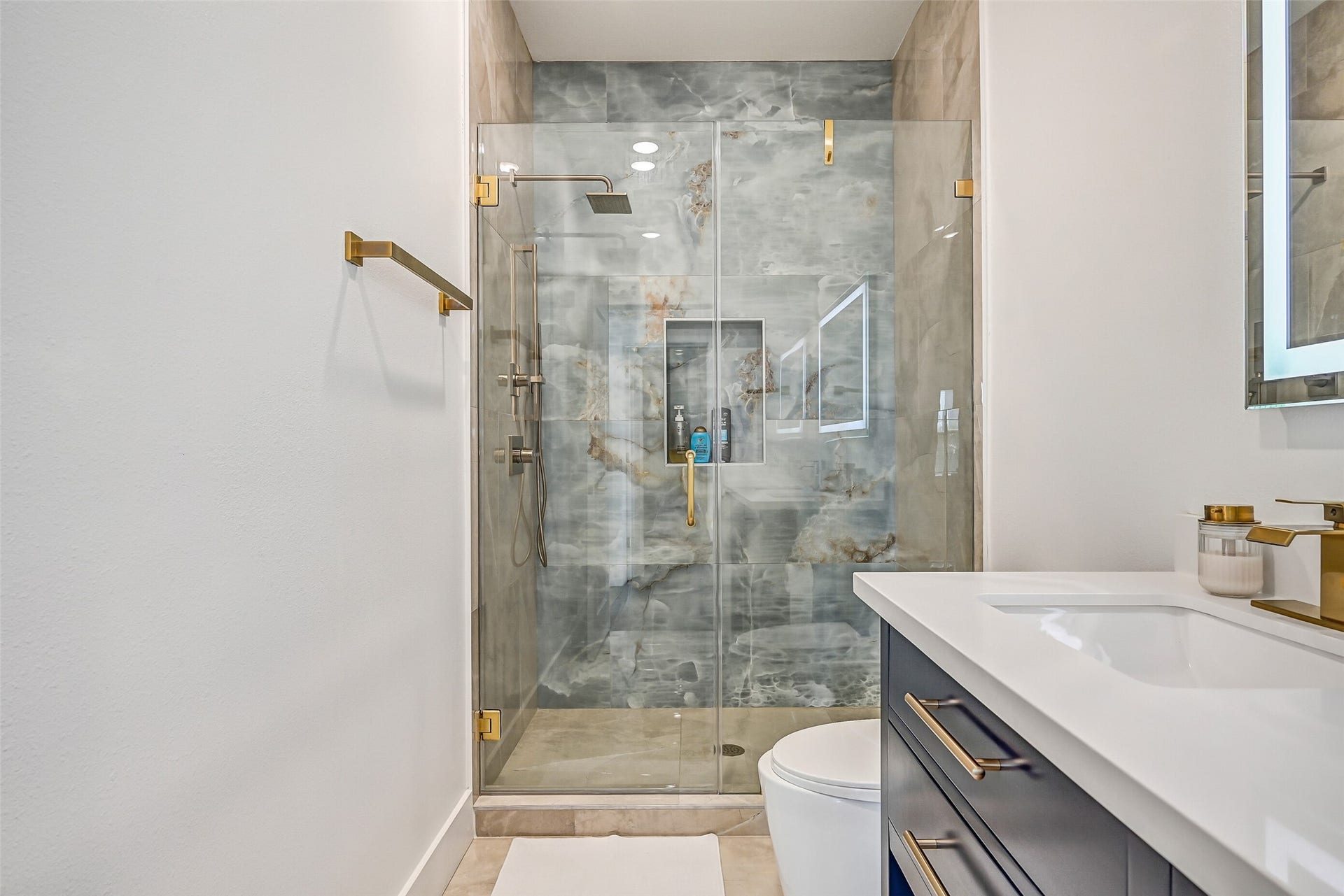3627 Niko Court, Galveston, TX 77554 This bathroom features quartz countertops, double sinks, gold fixtures, and a walk-in shower with a waterfall showerhead and stunning large tile surround.