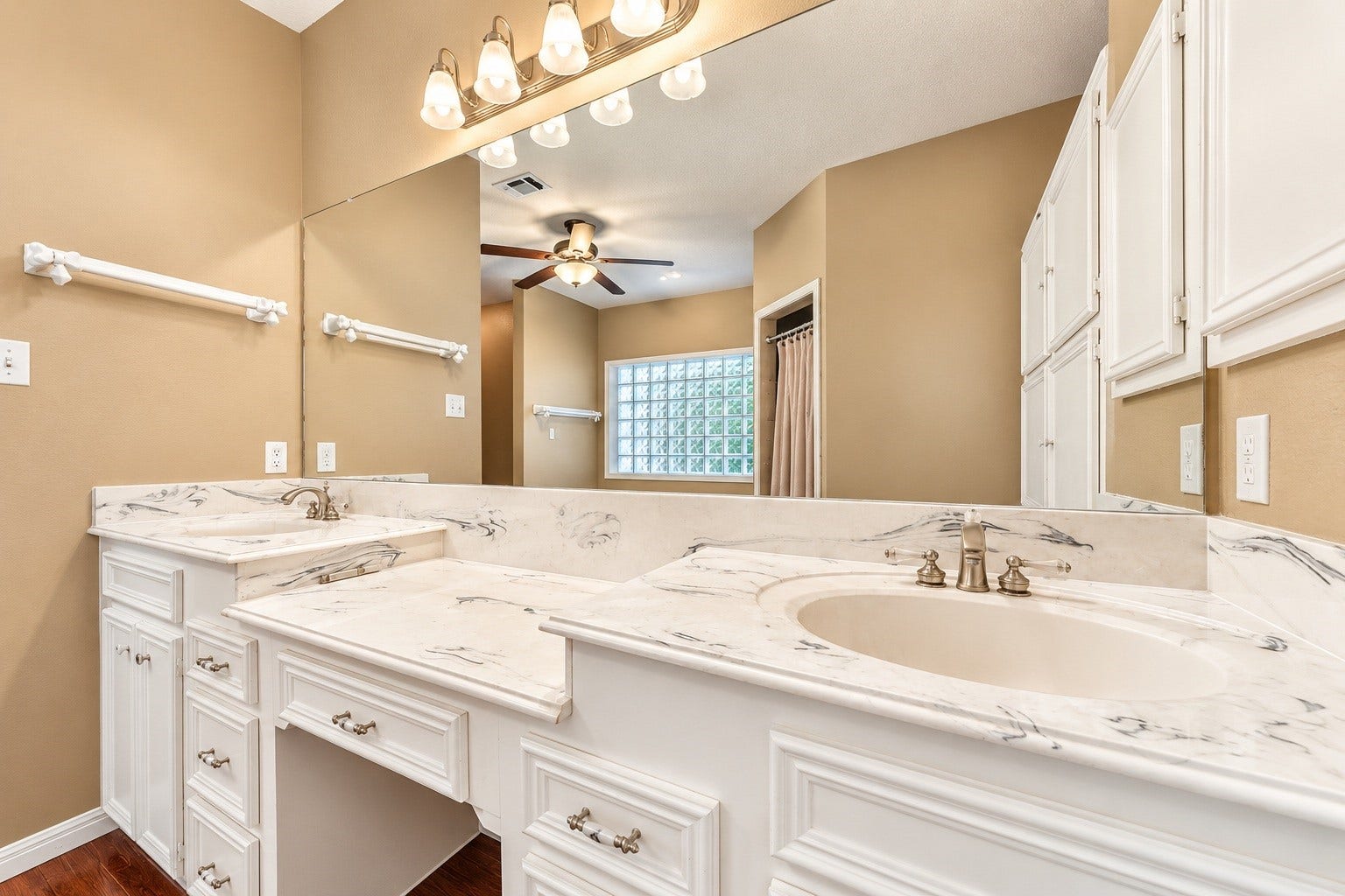 4030 Larkspur Street, Alvin, TX 77511 Elegant master bathroom featuring dual vanities with marble countertops, ample cabinet storage, and a large mirror, complemented by warm lighting and a ceiling fan.