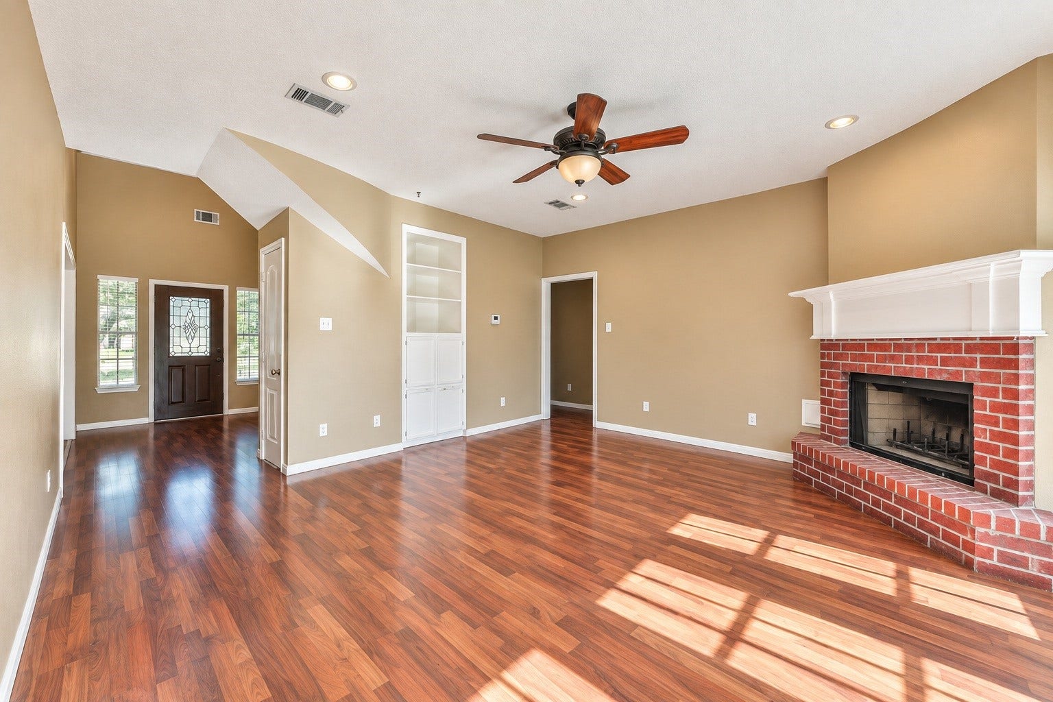 4030 Larkspur Street, Alvin, TX 77511 Spacious living room with rich wood-look floors, a cozy brick fireplace, and built-in shelving. Natural light pours in through large windows, complementing the ceiling fan and elegant entryway.