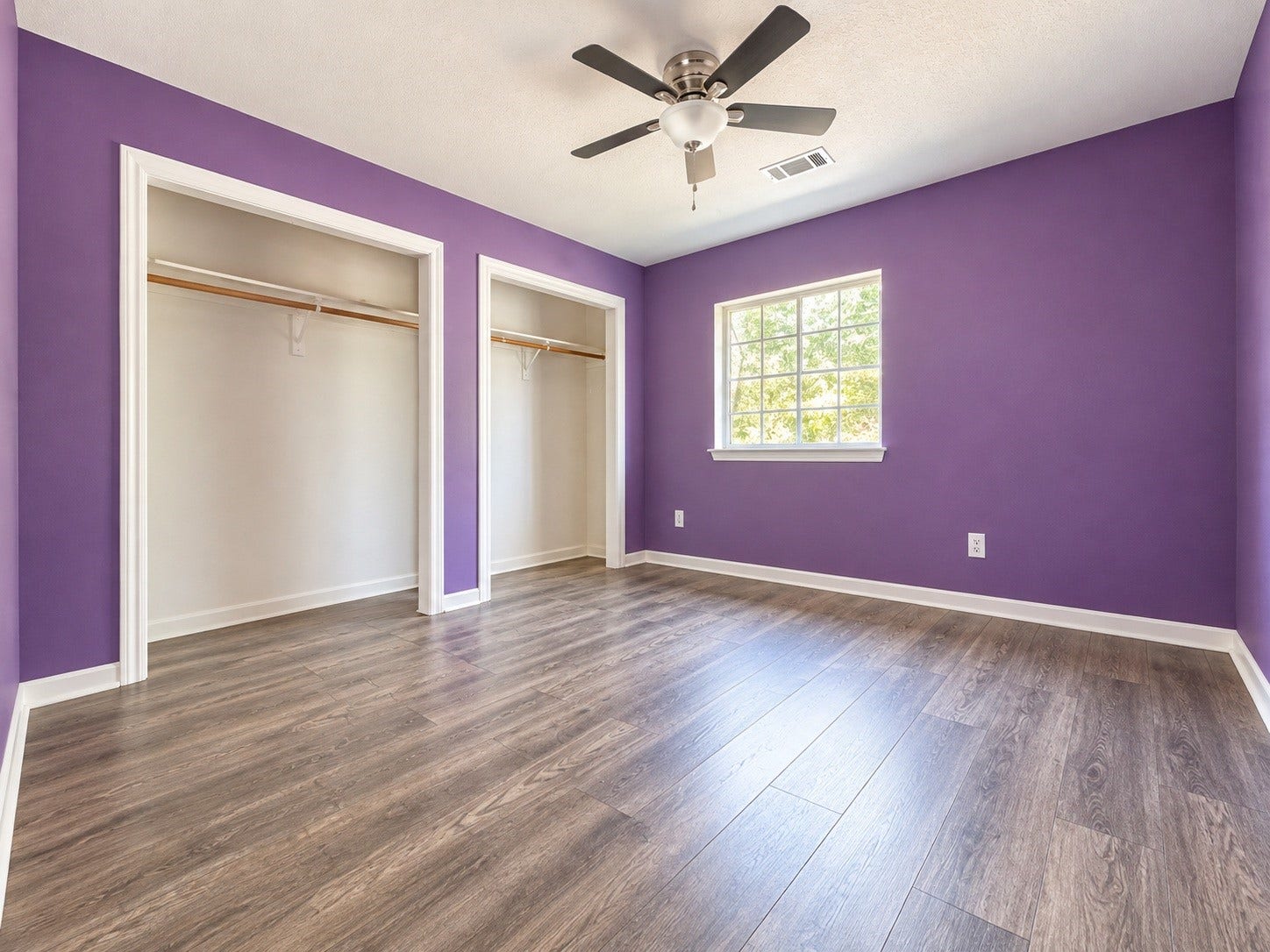 4030 Larkspur Street, Alvin, TX 77511 Bright bedroom with vibrant purple walls, dual closets, and warm wood-look flooring, complemented by a ceiling fan and a large window for natural light.