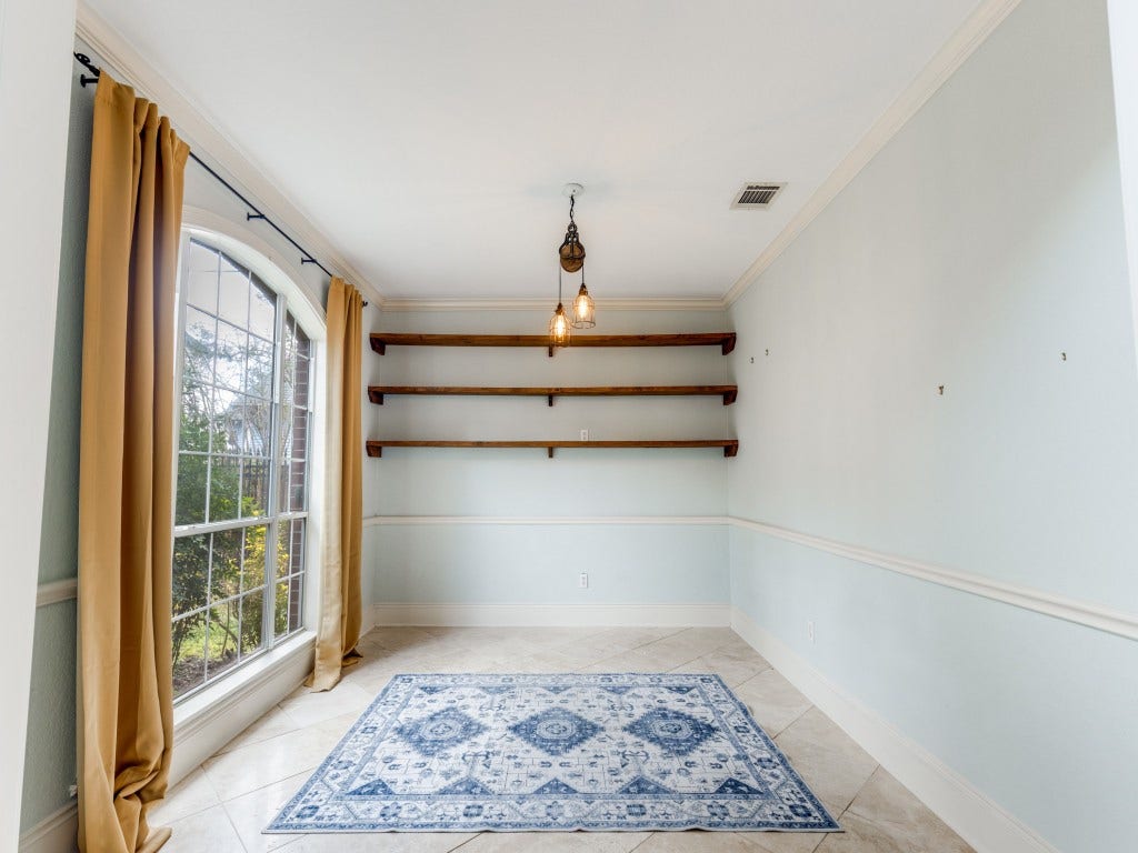 2811 Princeton Street, Houston, TX 77009 Bright room with large windows and elegant curtains, featuring three wooden shelves on a light blue accent wall. The space includes a stylish hanging light fixture and a patterned area rug on tiled flooring, perfect for a cozy reading nook or home office.