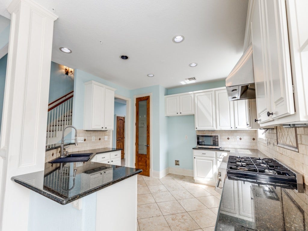2811 Princeton Street, Houston, TX 77009 This kitchen features modern white cabinetry, a gas stove, and granite countertops. It has a tiled backsplash, recessed lighting, and a bright, airy feel with light blue walls. The layout includes a breakfast bar and a view of the staircase, offering a spacious and inviting atmosphere.