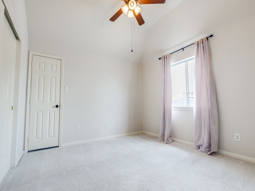 2811 Princeton Street, Houston, TX 77009 3rd bedroom on 2nd floor features a neutral color palette with plush carpeting, a ceiling fan, and ample natural light from the window with sheer curtains. A door leads to additional space or storage.