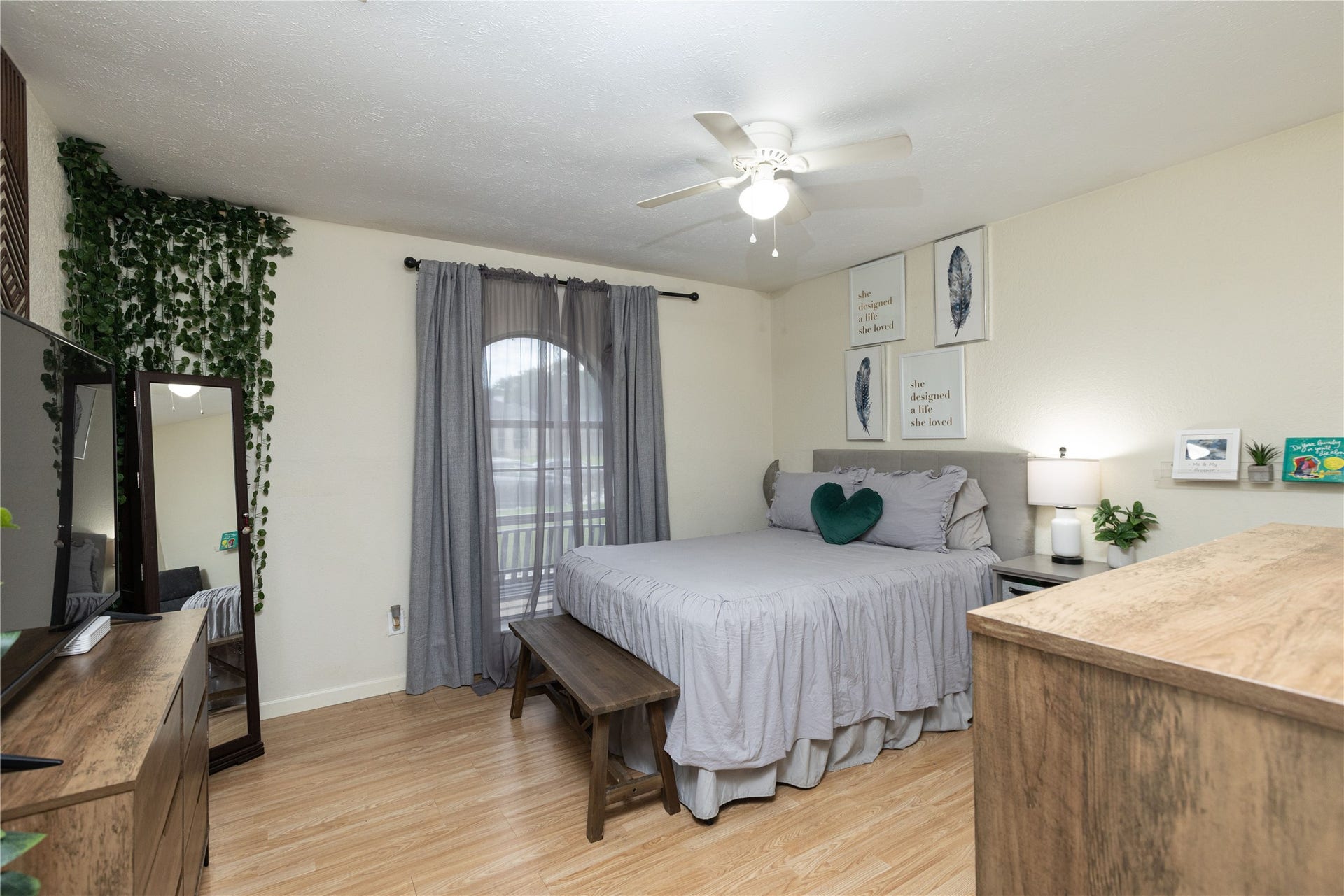 14007 Lorne Drive, Houston, TX 77049 Cozy bedroom with a calming neutral palette features a comfortable bed, elegant wall art, and a ceiling fan for added comfort. Natural light filters through sheer curtains, highlighting the warm wood flooring and inviting decor.
