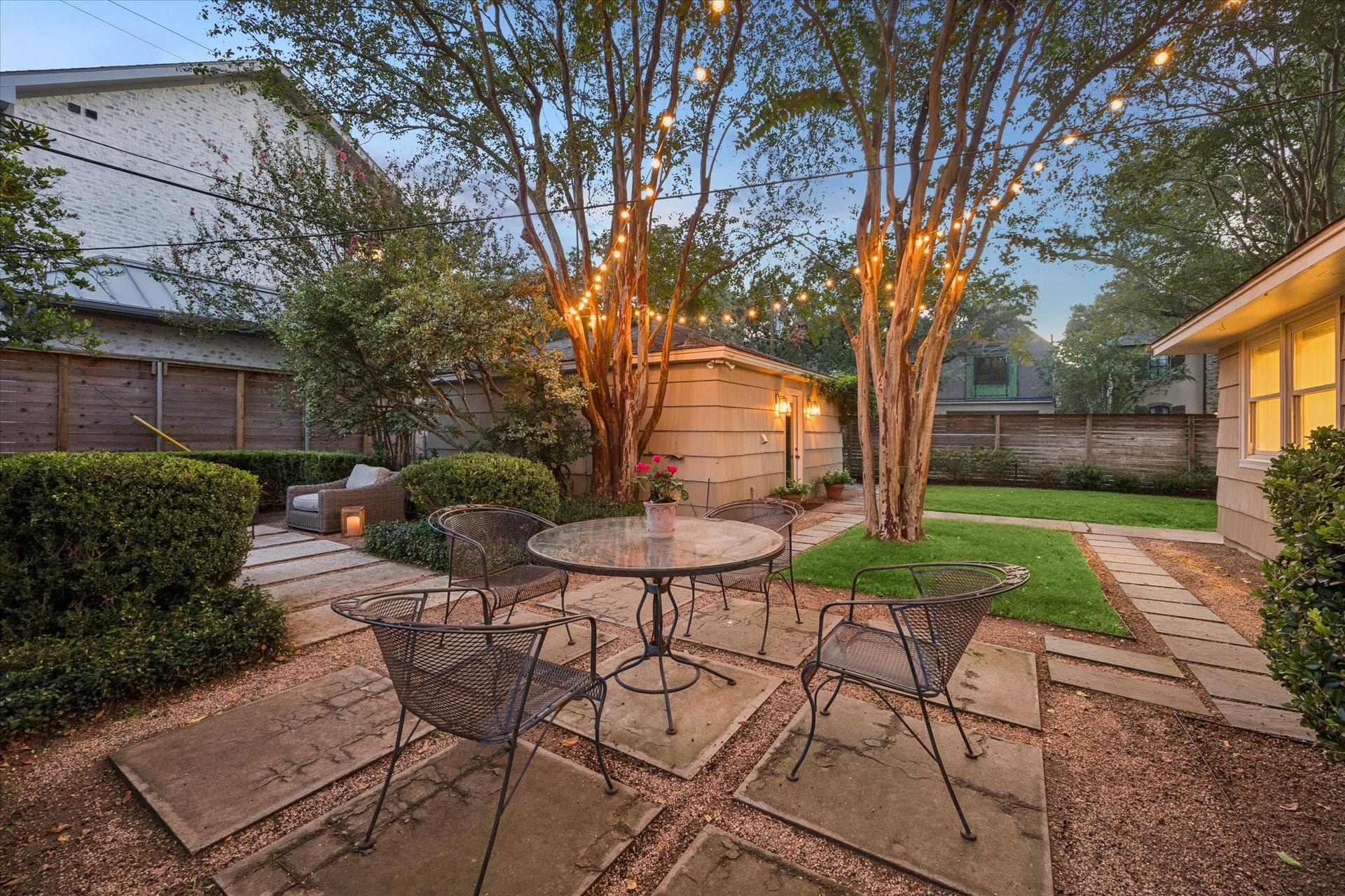 Charming backyard oasis featuring a cozy patio with string lights, surrounded by lush greenery and manicured landscaping, perfect for relaxing or entertaining.
