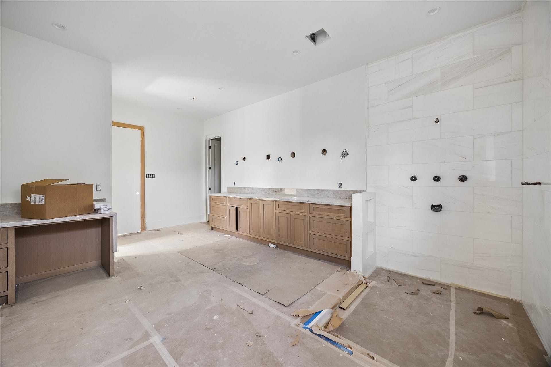 915 Lamonte Lane, Houston, TX 77018 Long vanity cabinetry combines drawers and enclosed storage beside a broad tiled bathing area. Natural tone wood and light veined tile establish the palette, while wall plumbing rough ins and protective floor coverings show the bath before fixtures and final fittings are installed.