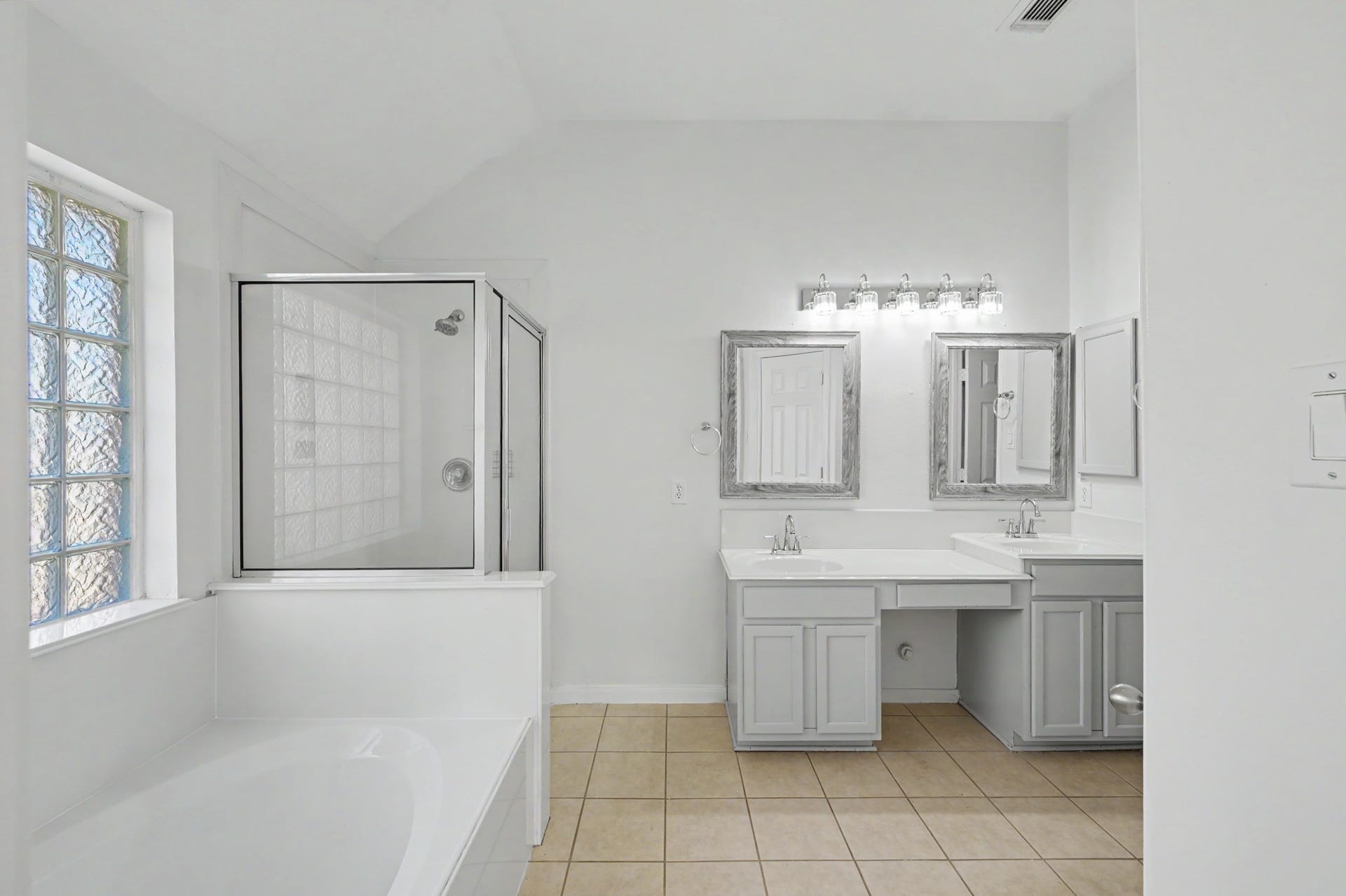 Spacious primary bath featuring dual vanities, soaking tub, separate glass-enclosed shower and decorative glass block window.