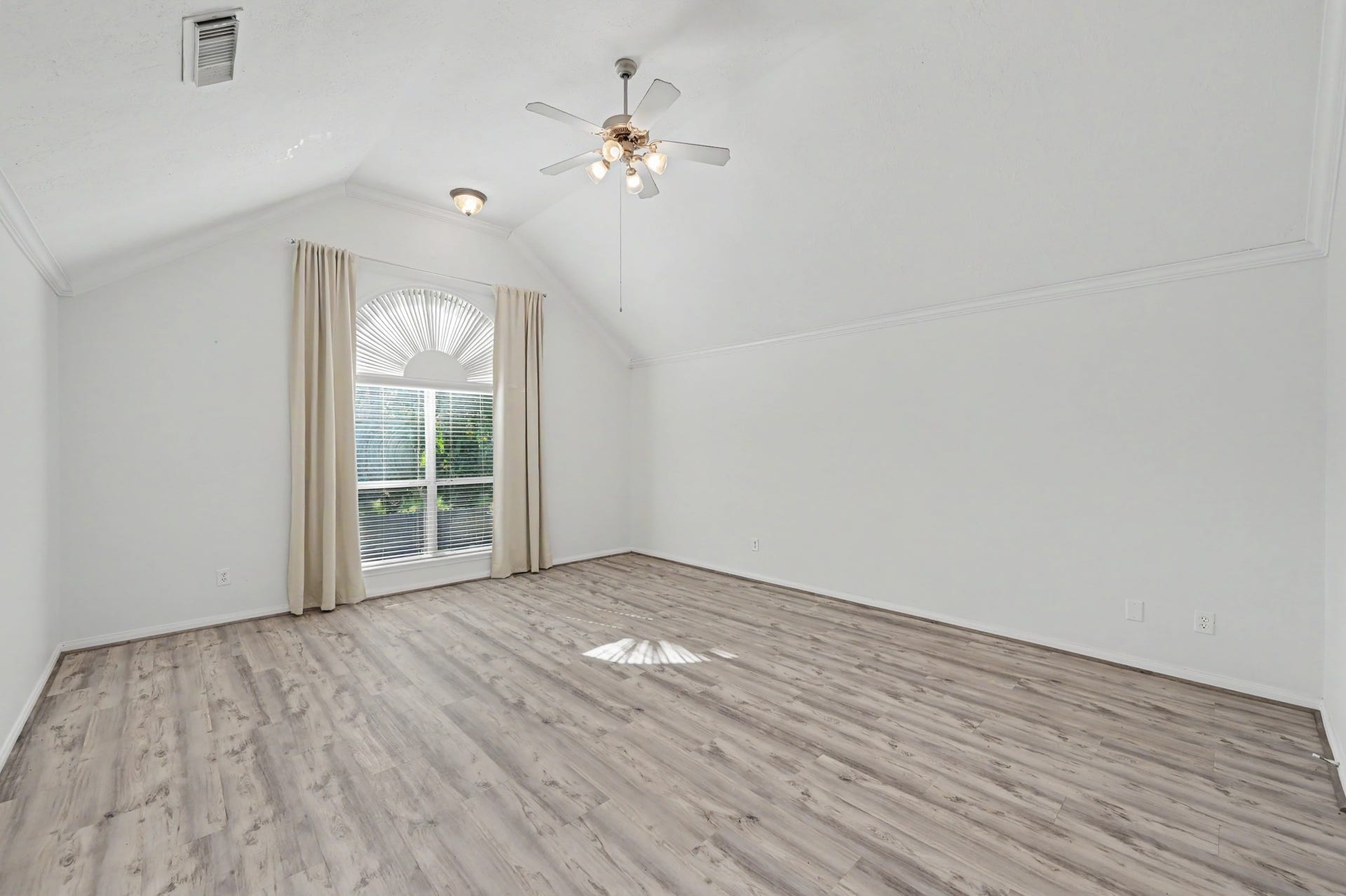 Oversized primary suite featuring vaulted ceilings, crown molding, wood-look flooring, ceiling fan and large arched window with abundant natural light.