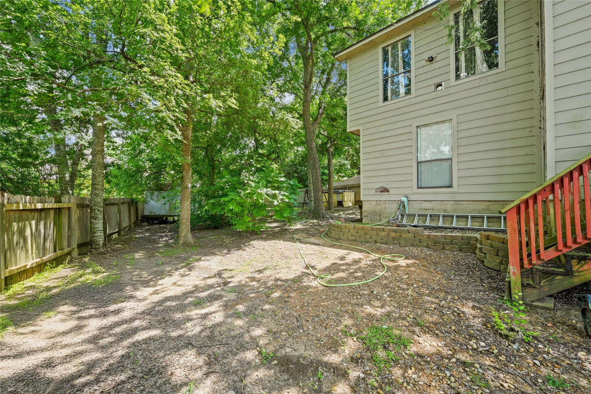 107 Harbour Town Lane, Conroe, TX 77356 A backyard view looking toward the side of a two-story beige house with a small wooden deck structure, surrounded by green trees and a wooden fence.