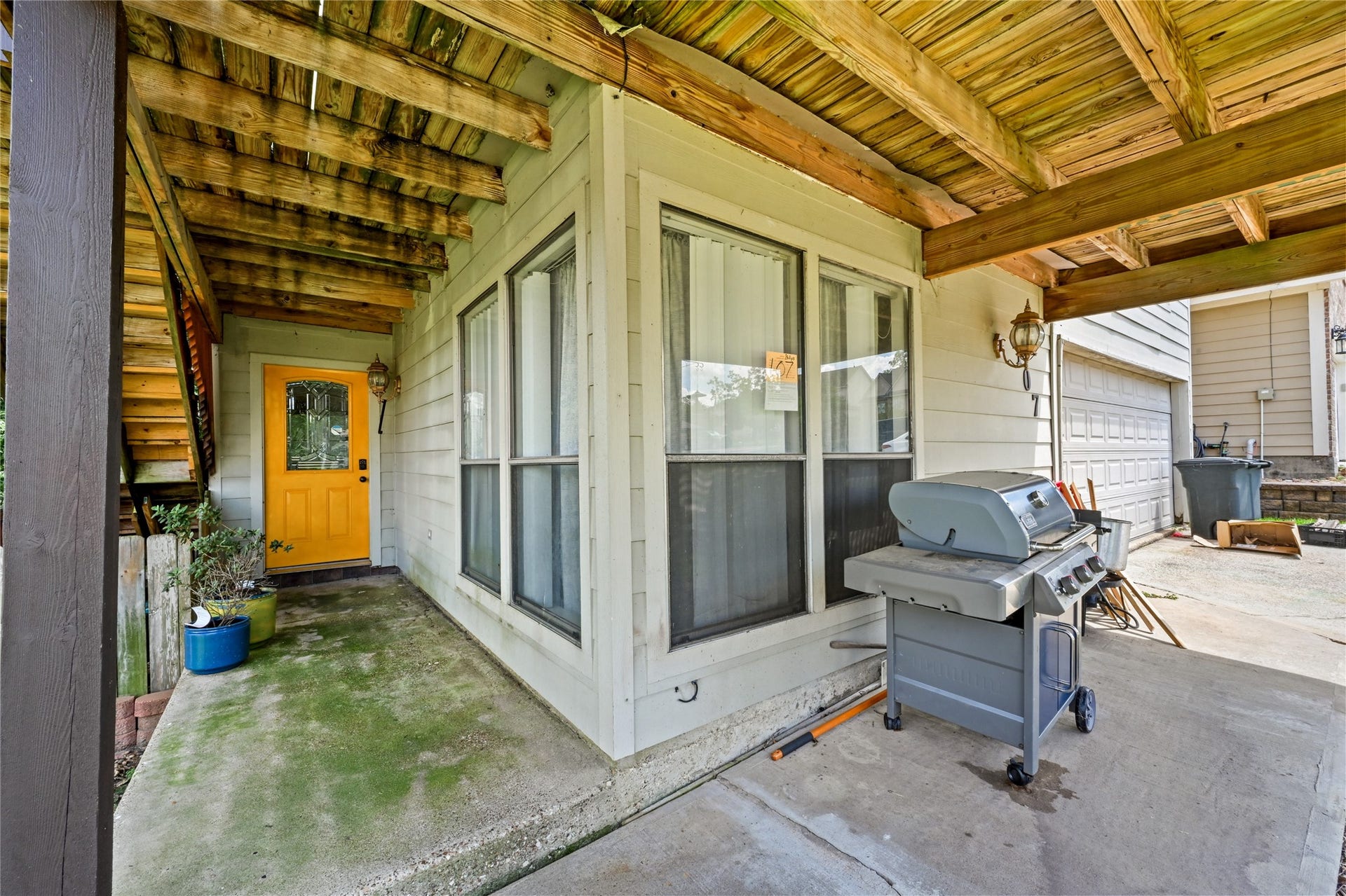 107 Harbour Town Lane, Conroe, TX 77356 A covered concrete patio space beneath an upper deck, featuring large windows, a entry door, and a barbecue grill stationed outside.