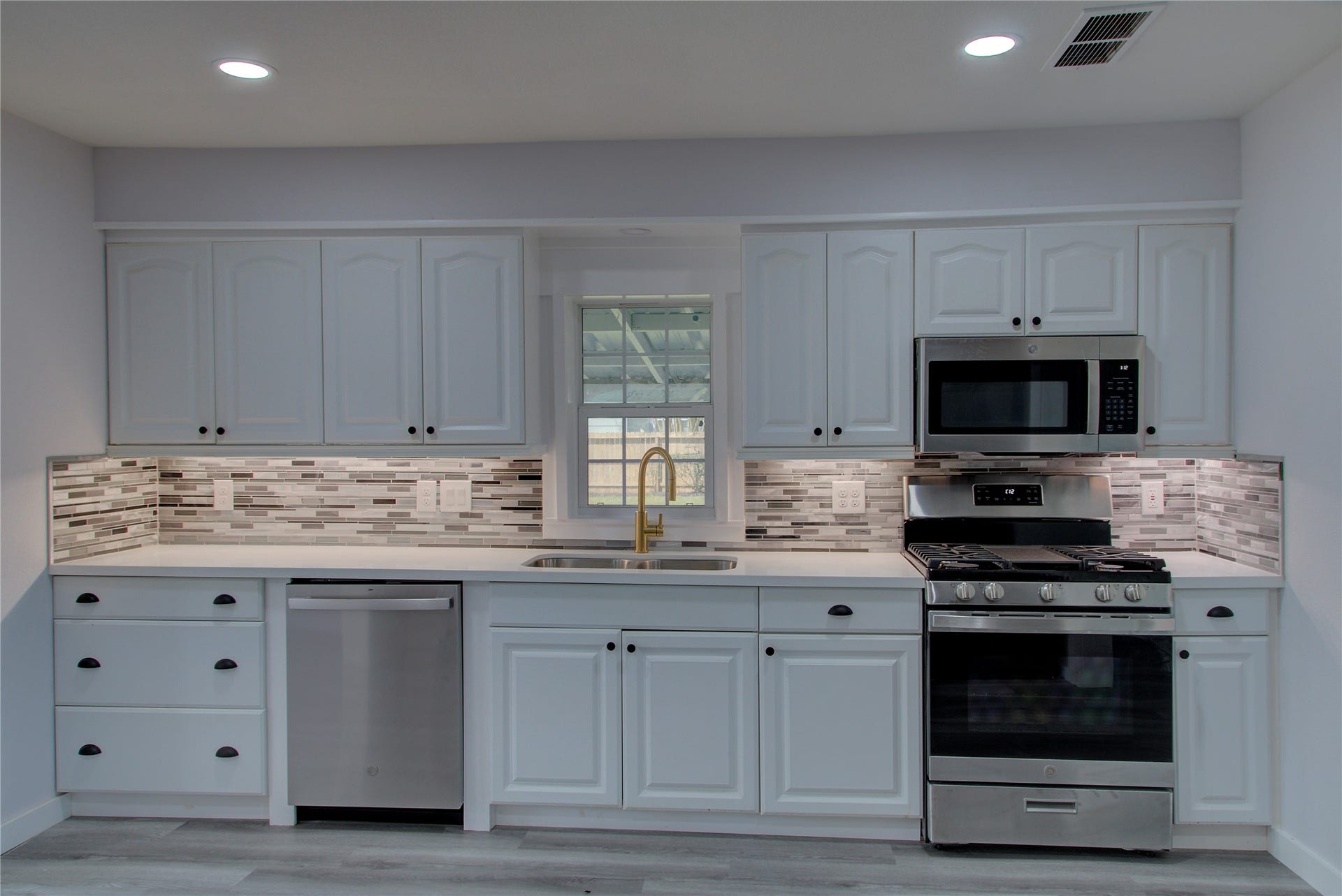 415 Perkins Avenue, League City, TX 77573 This modern kitchen features sleek white cabinetry, stainless steel appliances, and a stylish tile backsplash. It includes a gas stove, microwave, dishwasher, and a window above the sink, providing ample natural light.