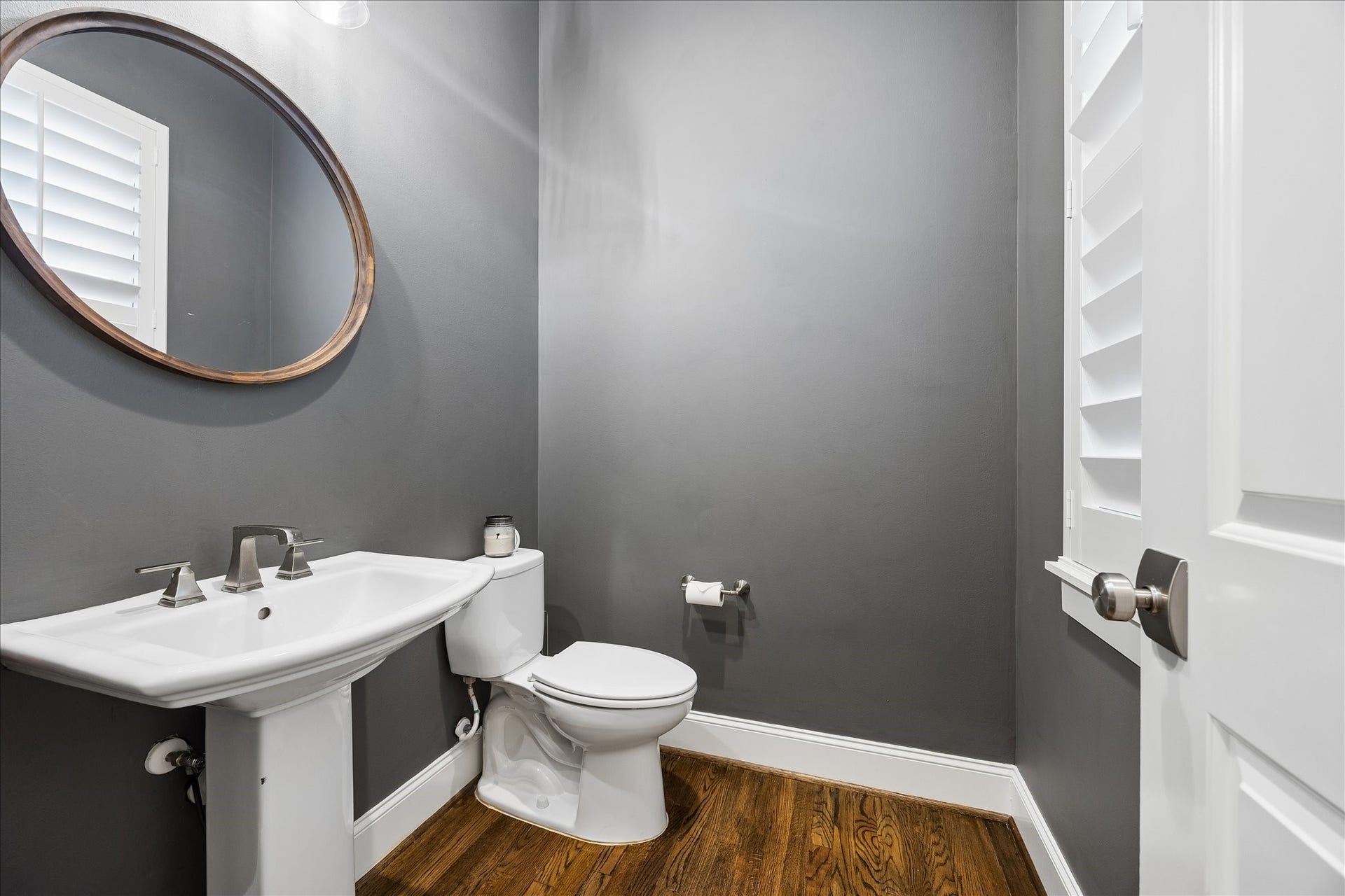 2915 Hicks Street, Houston, TX 77007 A stylish powder room features sleek gray walls, a modern pedestal sink, and a round mirror, complemented by rich wood flooring and natural light from a shuttered window.