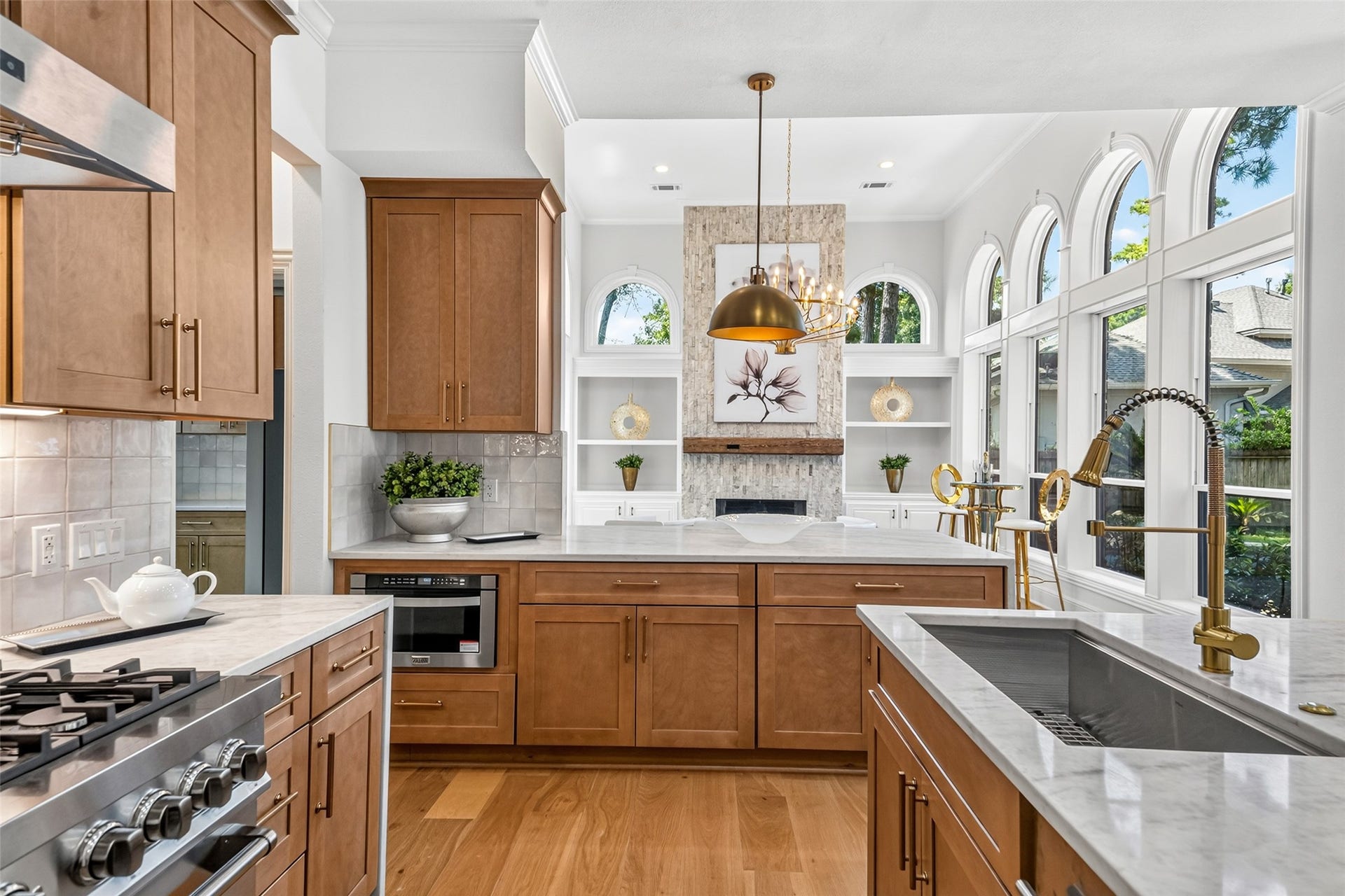 46 Pebble Cove Drive, The Woodlands, TX 77381 Elegant kitchen featuring warm wood cabinetry, sleek marble countertops, and a modern gas range. Expansive arched windows flood the space with natural light, highlighting the stylish pendant lighting and chic fixtures.