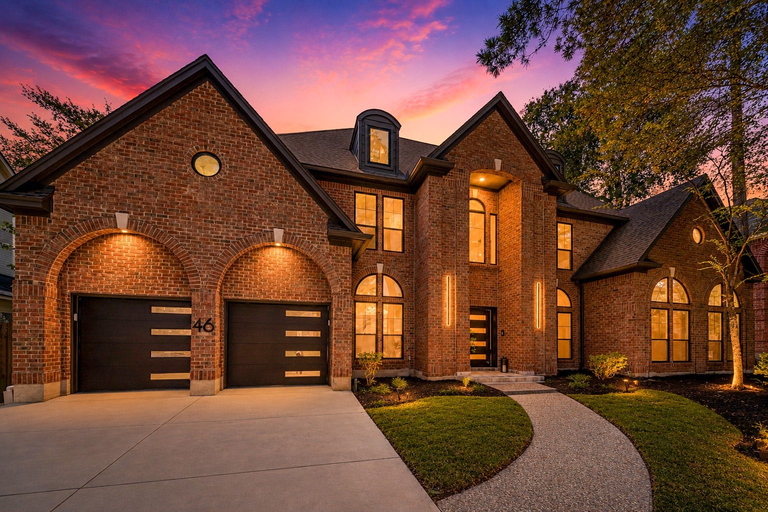 46 Pebble Cove Drive, The Woodlands, TX 77381 Stunning brick home with elegant arched windows, modern garage doors, and ambient exterior lighting, set against a picturesque sunset backdrop.