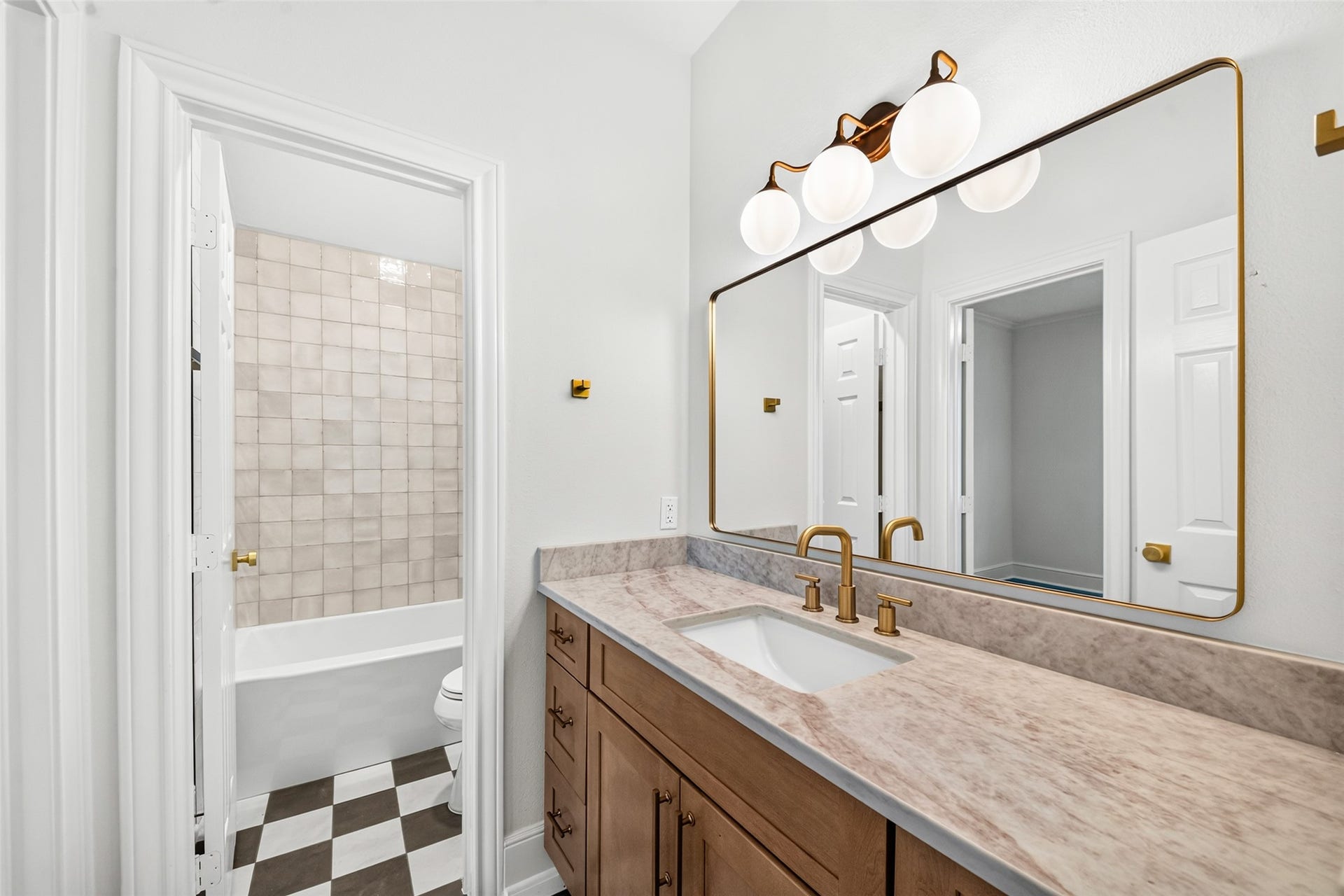 46 Pebble Cove Drive, The Woodlands, TX 77381 Elegant bathroom featuring a stylish vanity with a marble countertop, brass fixtures, and a large framed mirror. The adjacent bathtub area showcases classic tilework, complemented by a chic black-and-white checkered floor.